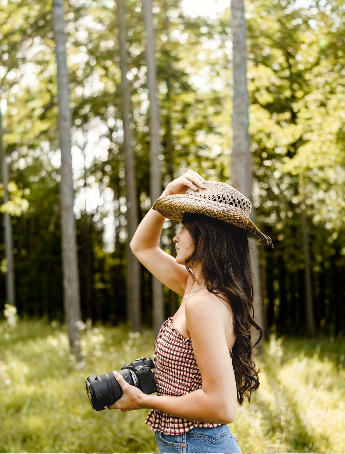 A young woman in a straw hat and checkered top holds a camera in a sunlit forest clearing.