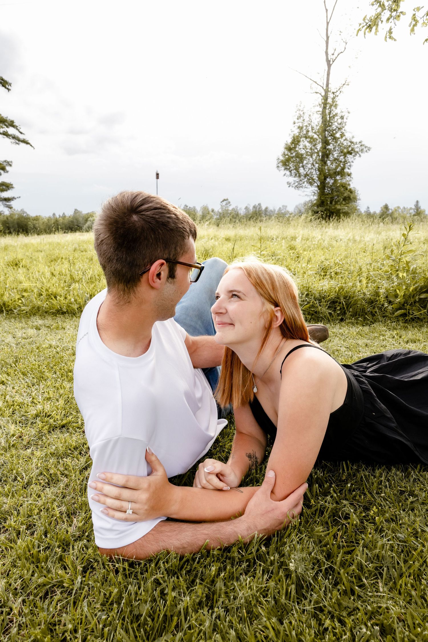 A couple relaxes on green grass in a park, smiling at each other in a tender romantic moment outdoors.