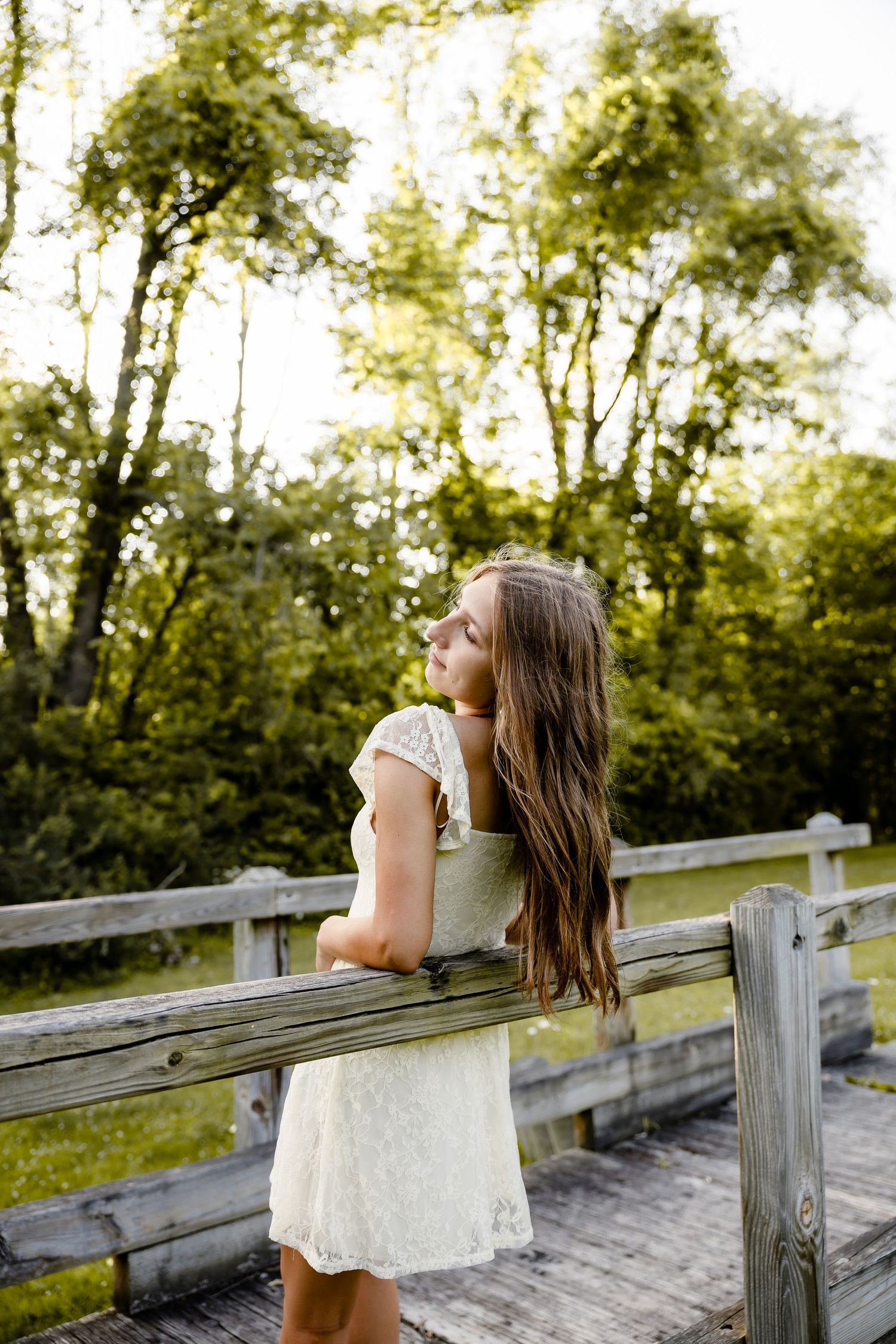 Young woman in white lace dress leaning on wooden fence, surrounded by lush green trees in a serene outdoor setting.