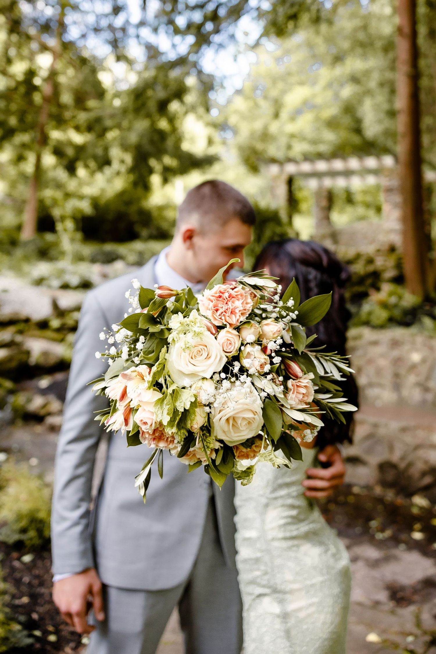 A bride and groom kissing behind a lush bridal bouquet of white and peach roses in a garden setting.