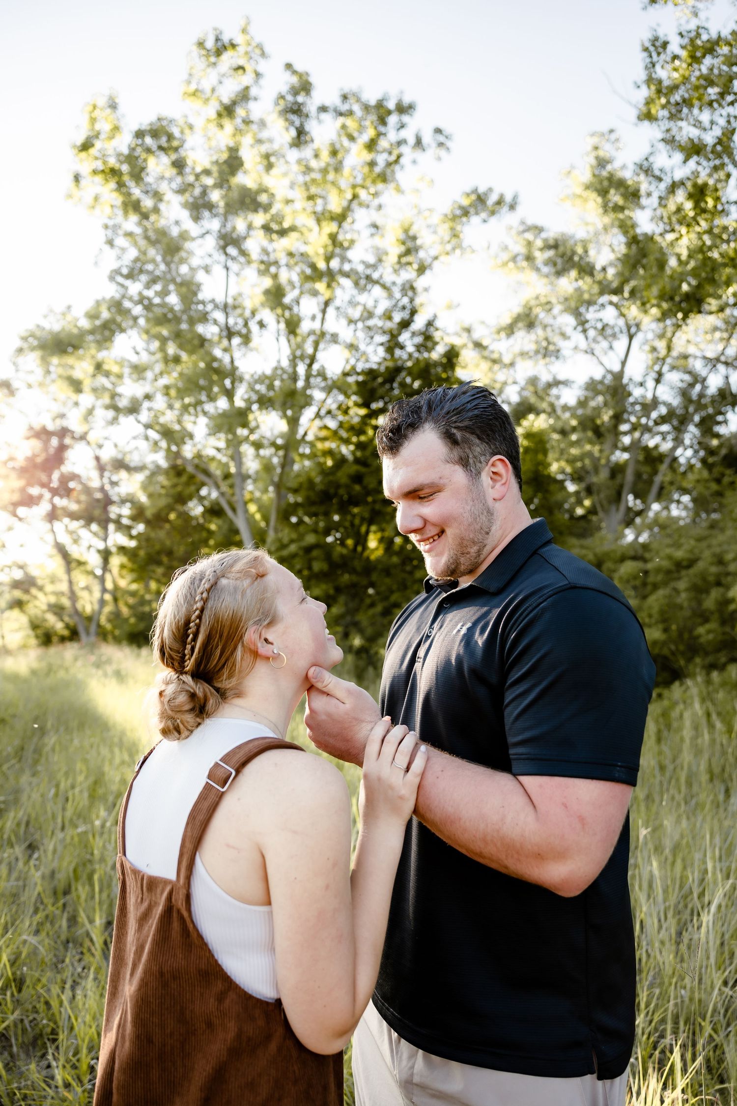 Couple sharing a tender moment in a sunlit field, man in black polo gently touching woman's face outdoors.