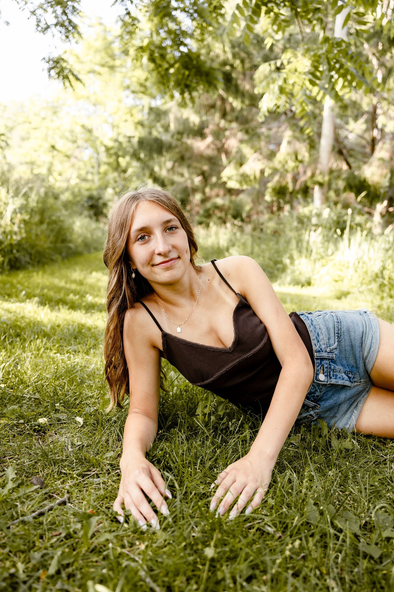 Young woman in black tank top and denim shorts relaxing on green grass in a sunny park setting.