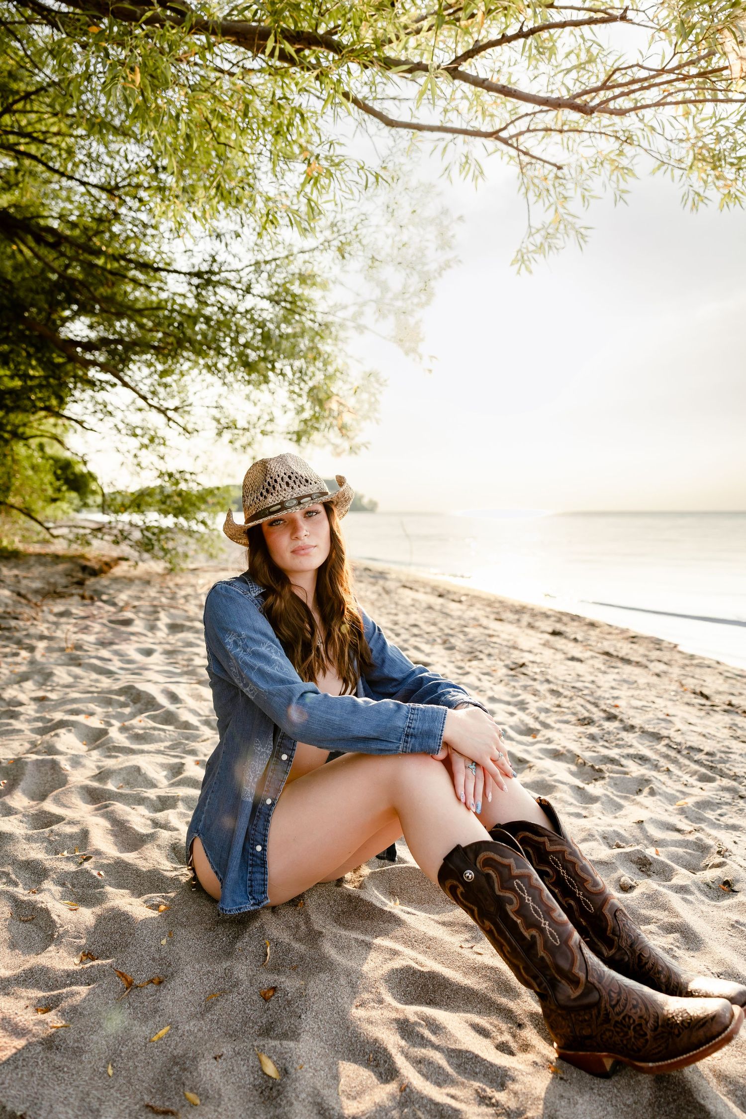 Young woman in cowboy hat, denim shirt, and boots sitting on sandy beach under trees near calm water.