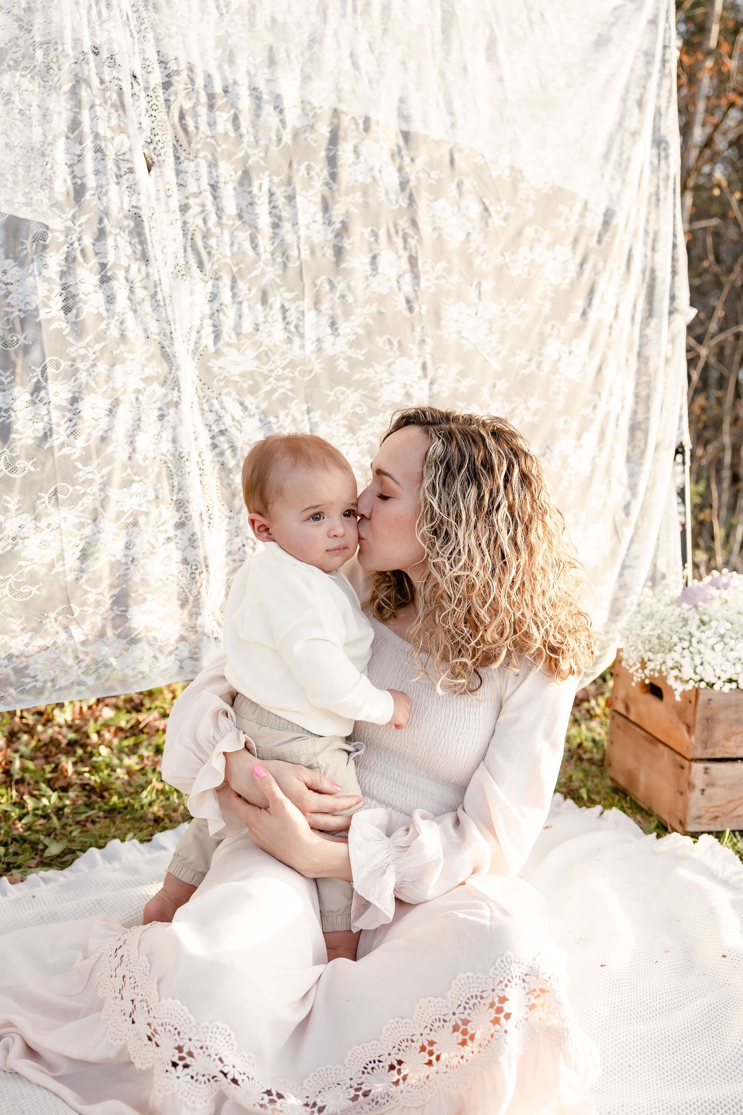 Curly-haired mother in white dress kissing her baby outdoors against a lace backdrop.