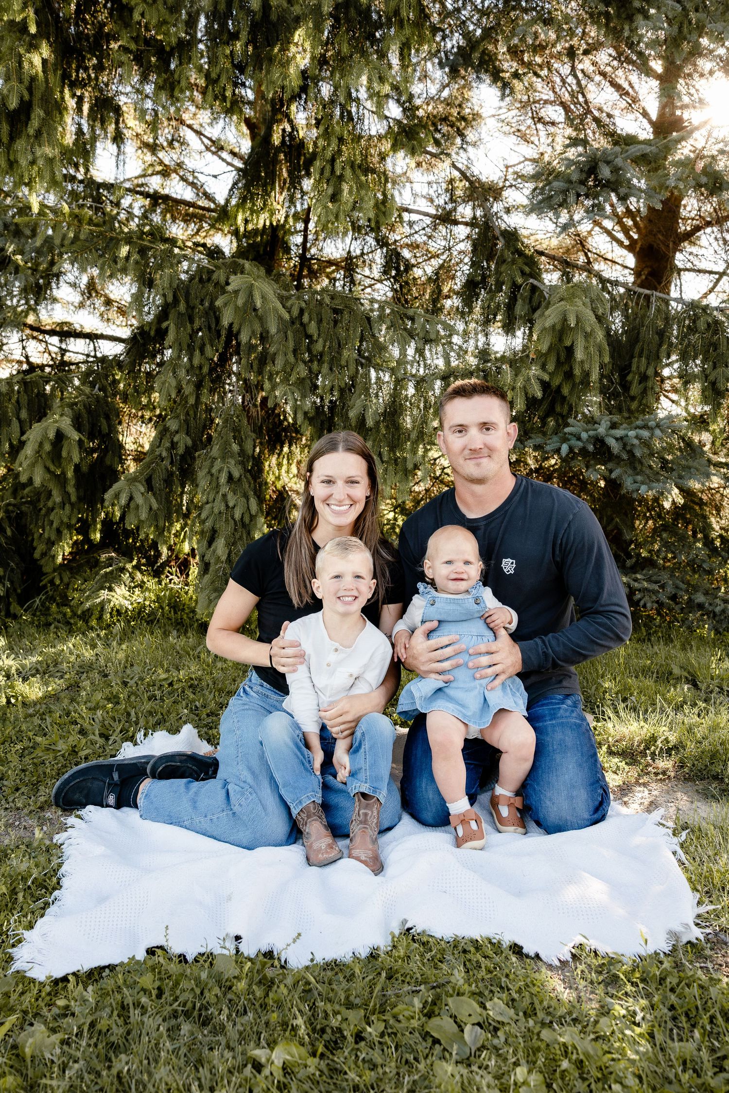 Family of four smiling outdoors on white blanket under pine trees, parents holding two young children in natural light.