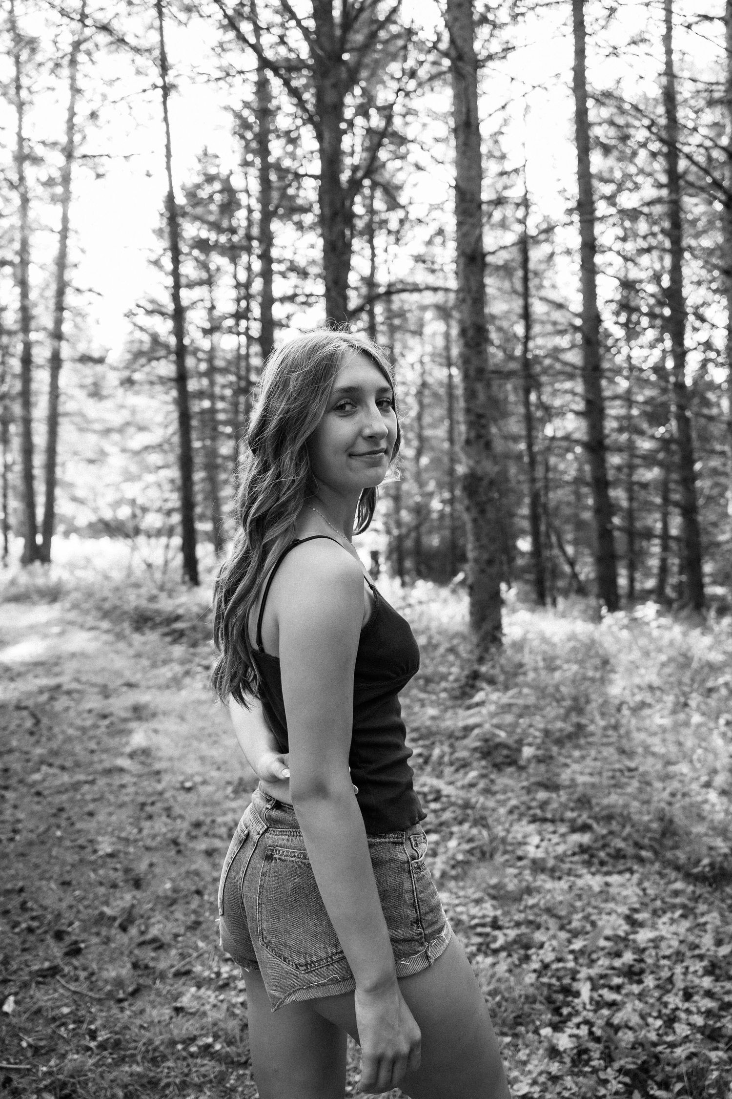 Black and white photo of a young woman in denim shorts posing in a forest with tall trees in the background.