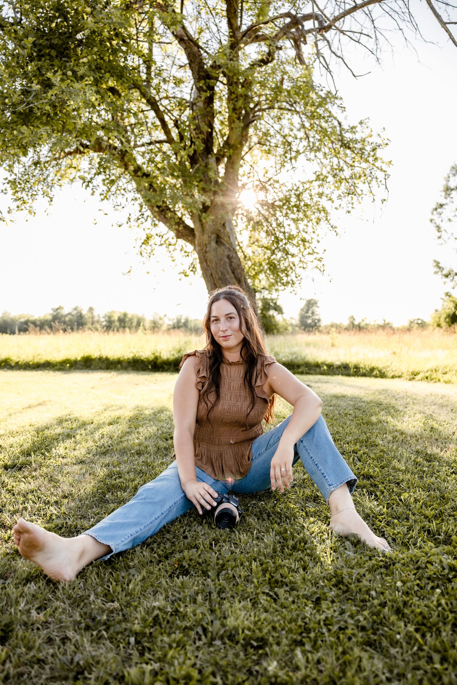 Woman sitting barefoot on grass under a sunlit tree, holding a camera, wearing brown top and jeans.