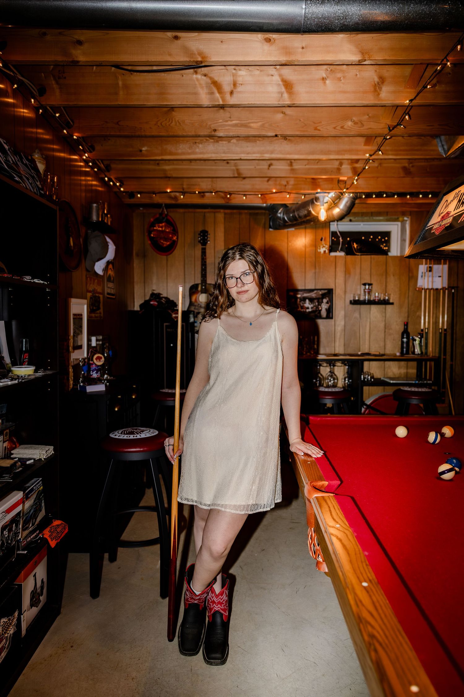 Woman in white slip dress holding pool cue beside red billiard table in cozy wood-paneled game room.