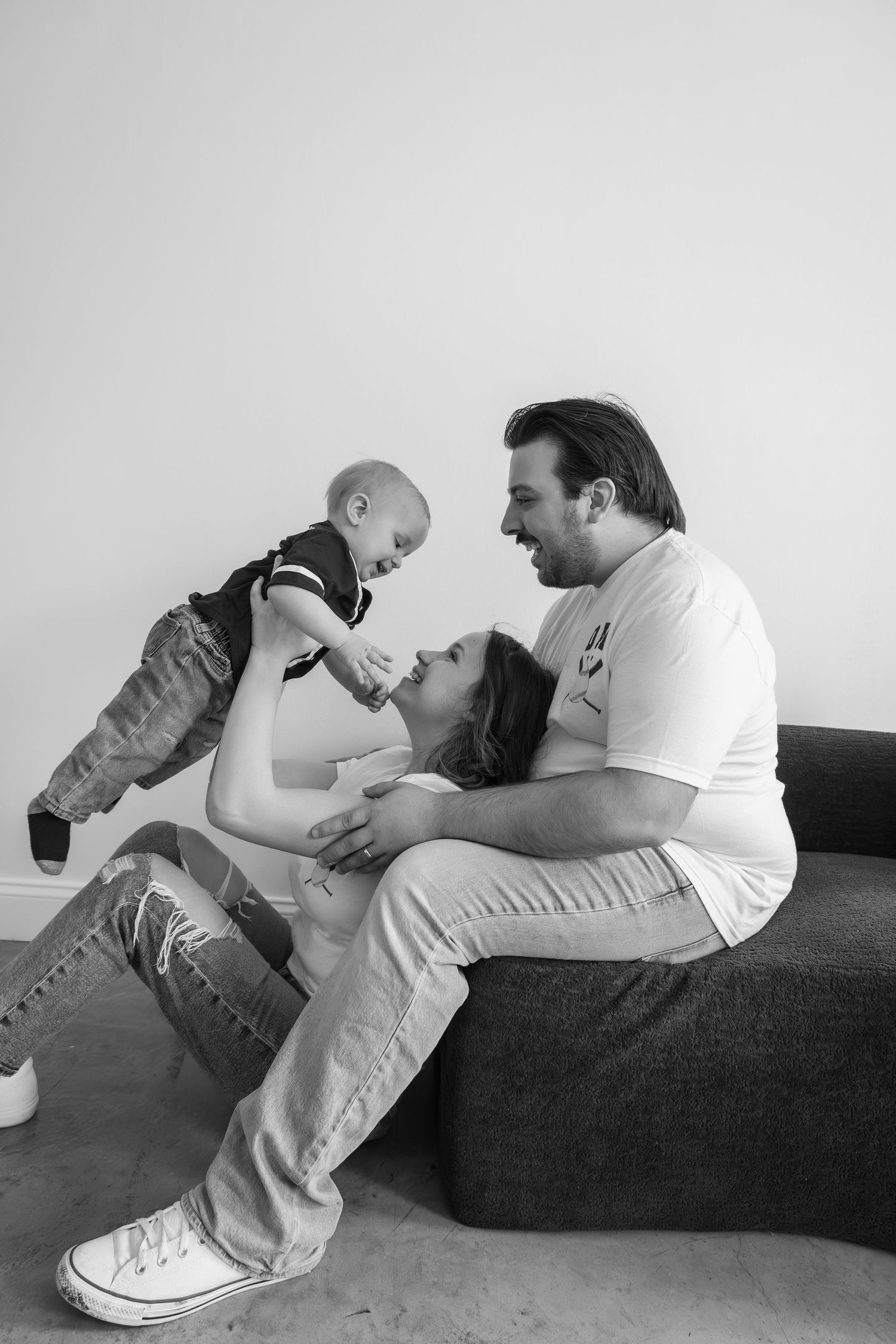 Black and white photo of a happy family with a pregnant woman, man, and baby sitting together on a couch.