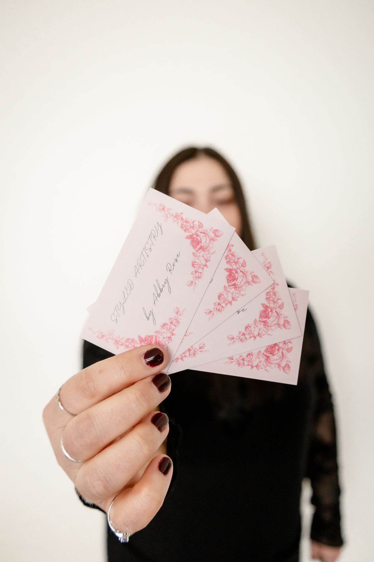 Woman in black dress holding pink floral wedding invitations fanned out toward camera with dark manicured nails.
