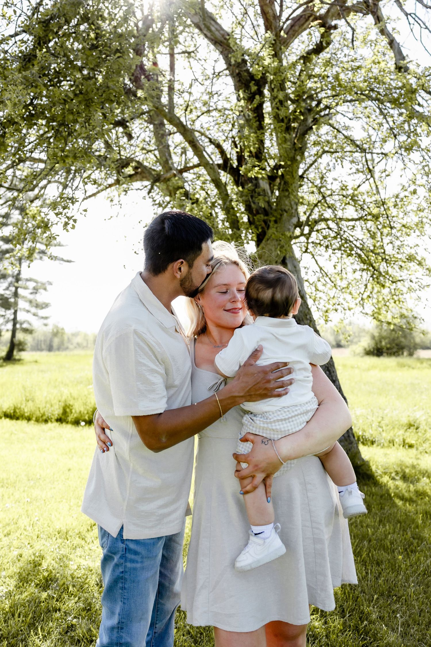 A couple shares a tender moment outdoors, as the man kisses the woman holding their baby near a blooming tree.