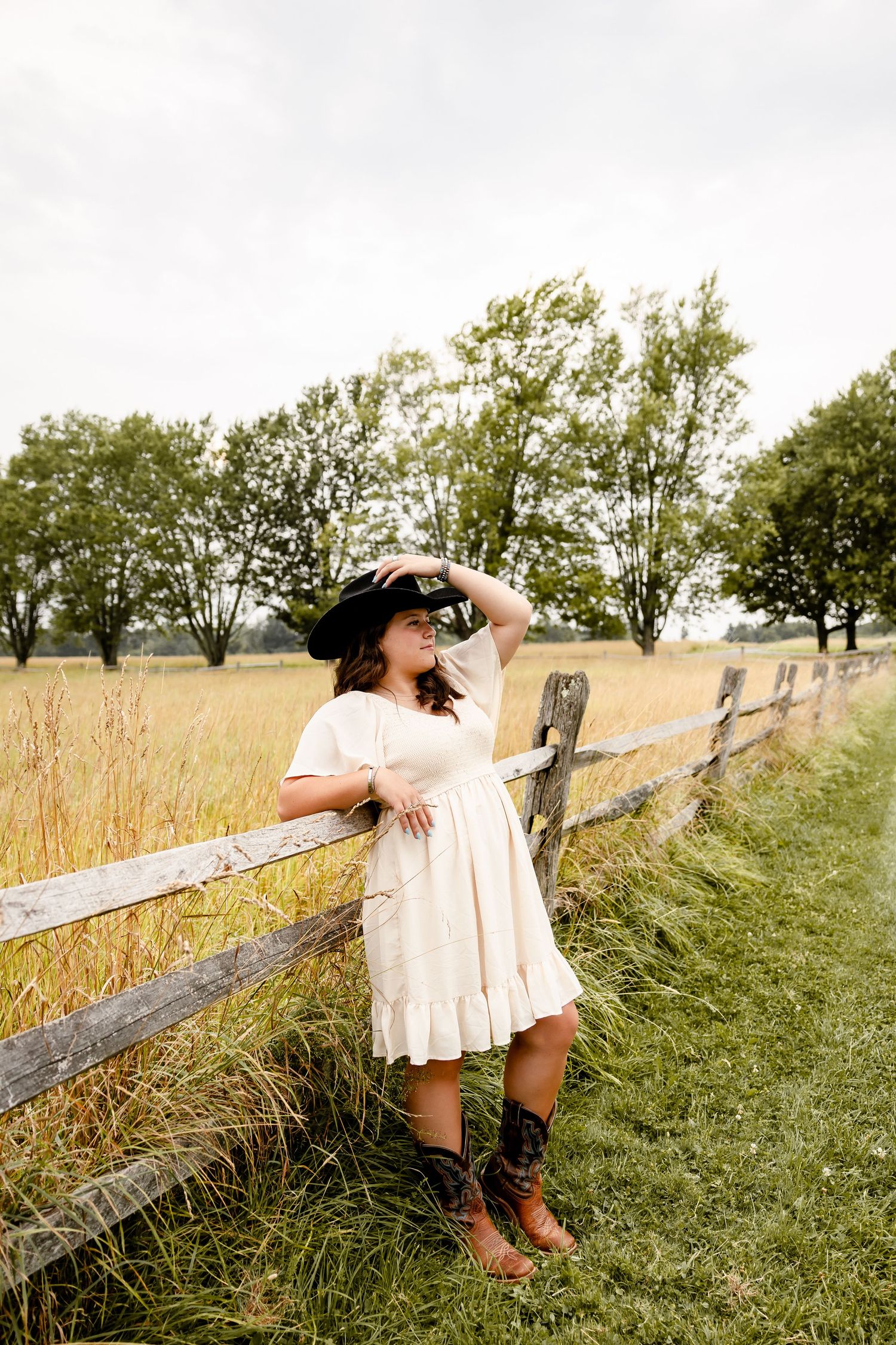 A young girl in a white dress, cowboy hat, and boots leans on a rustic wooden fence in a golden field.