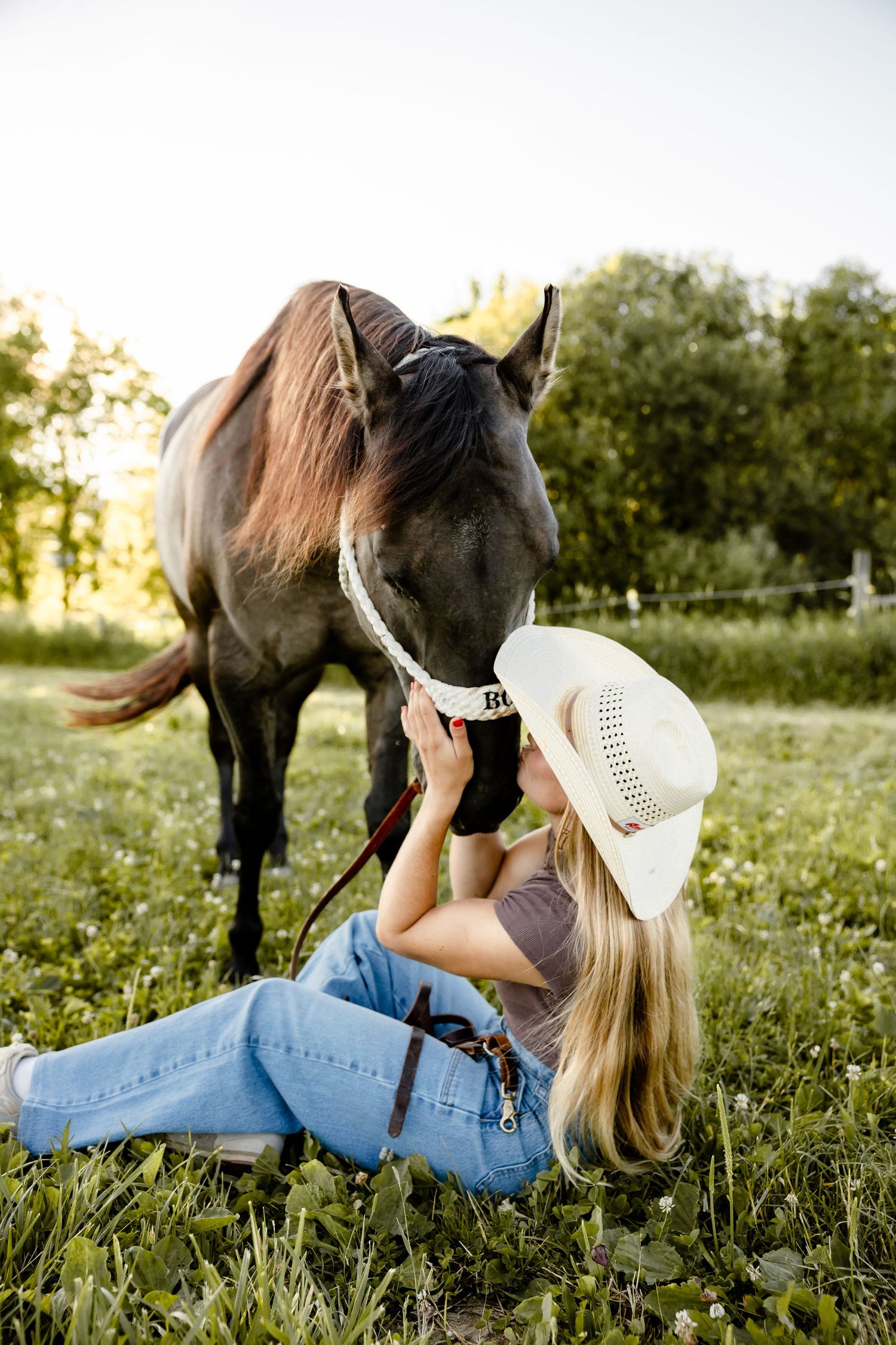 Blonde woman in cowboy hat sitting on grass kissing a dark horse on a sunny day in a green field.