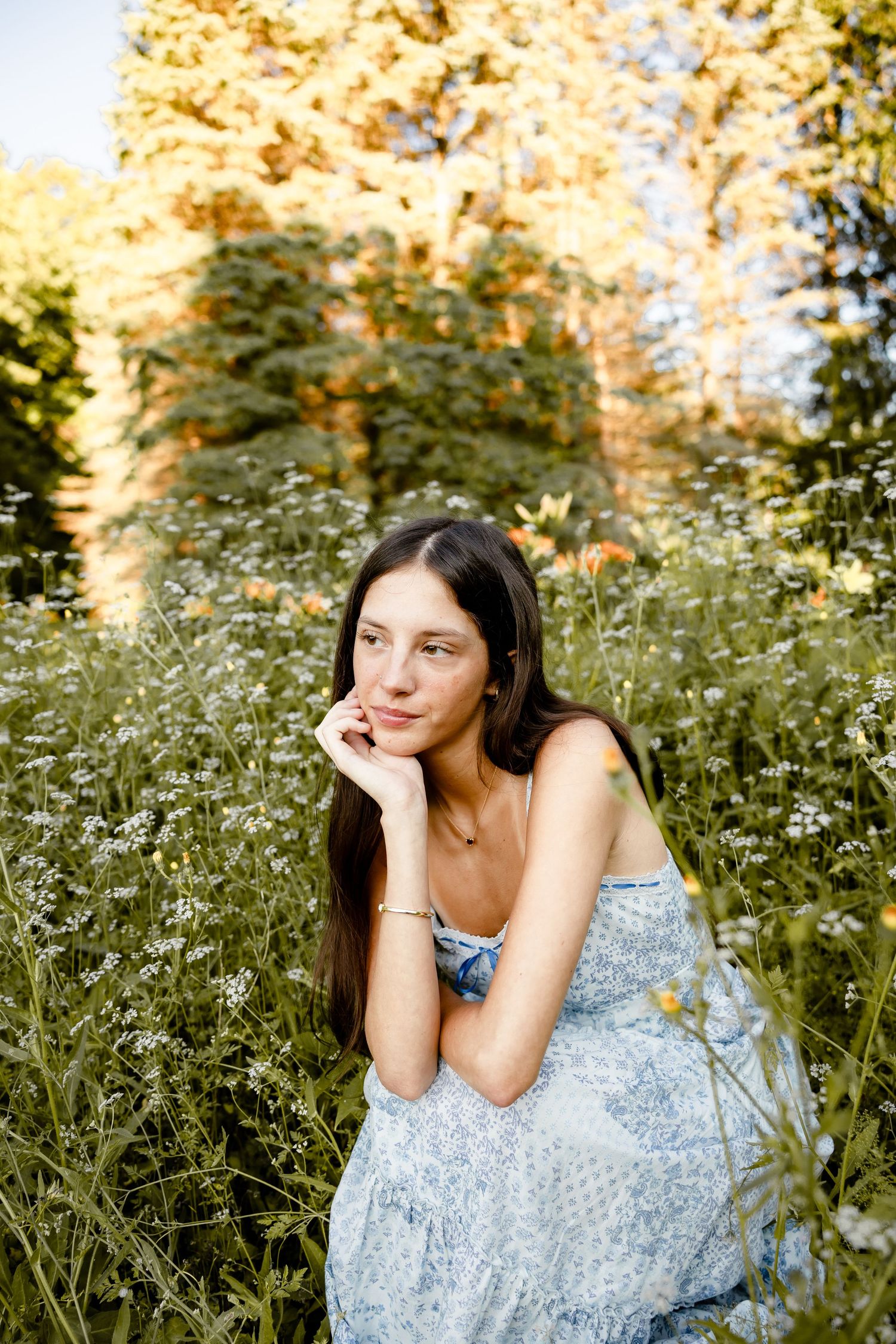Young woman in blue floral dress sitting among wildflowers in a sunlit garden, resting chin on hand.