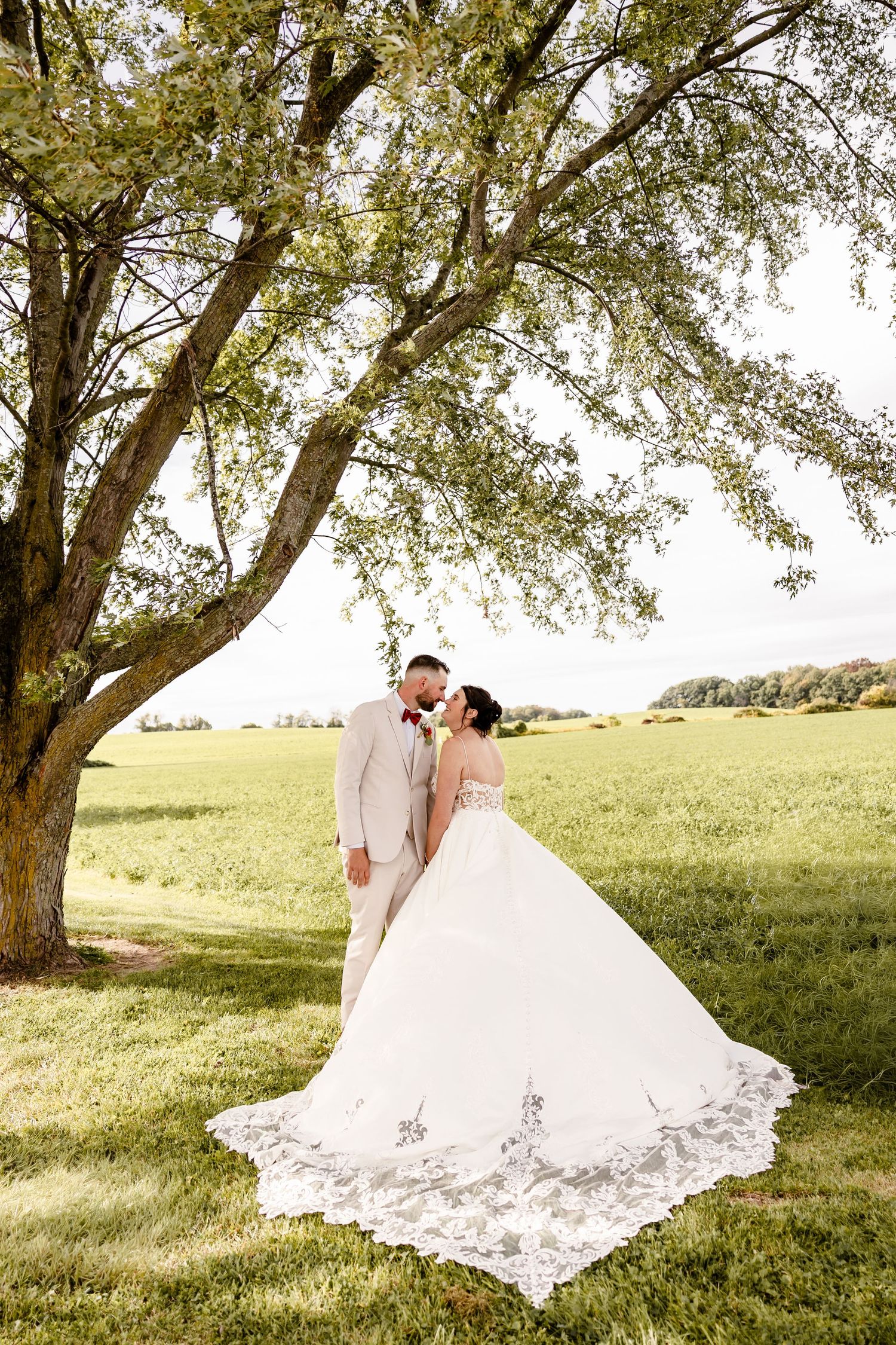 Bride and groom share a romantic moment under a large tree in a lush green field on their wedding day.