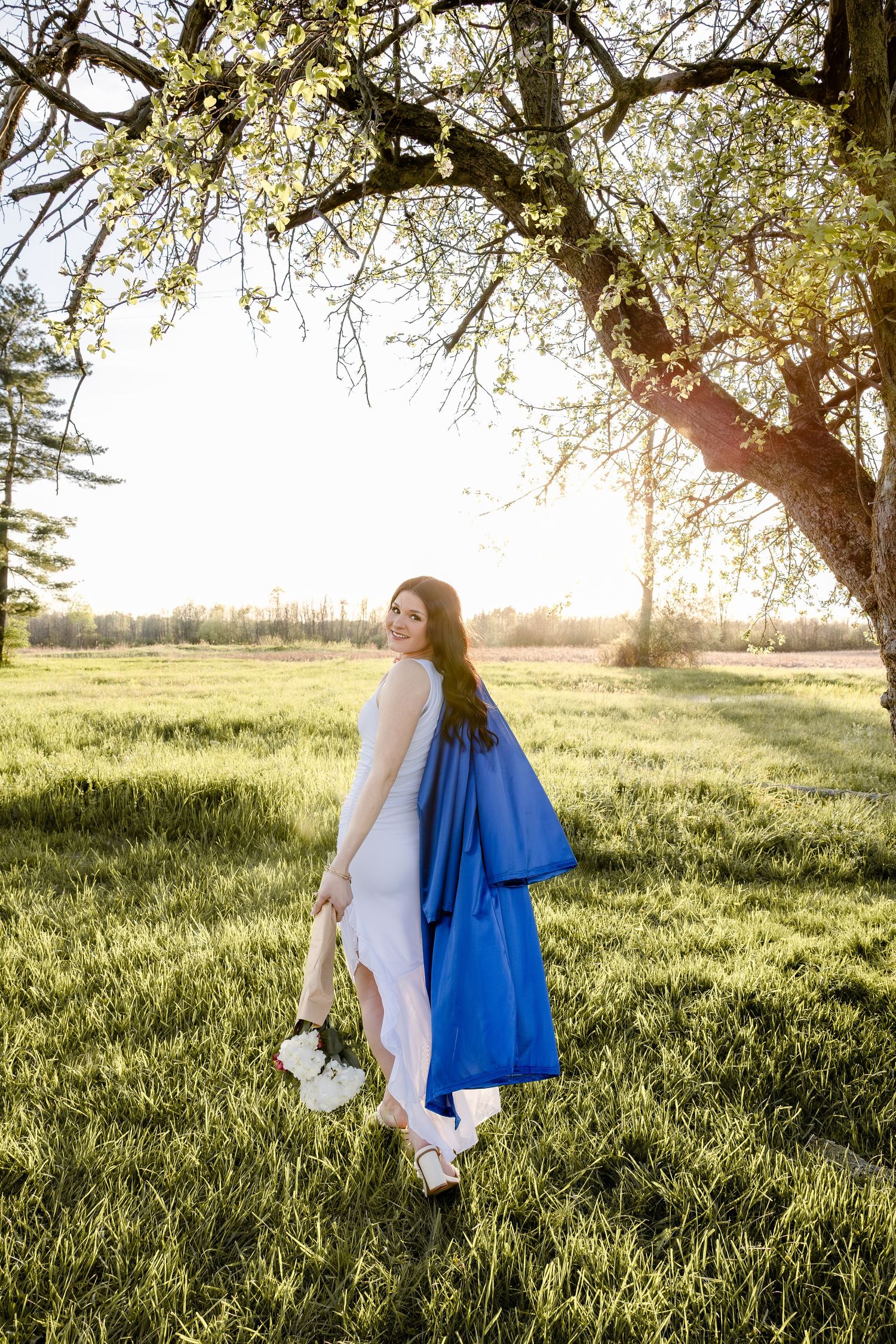 Young woman in white outfit holding blue graduation gown, standing under blooming tree in sunny green field.