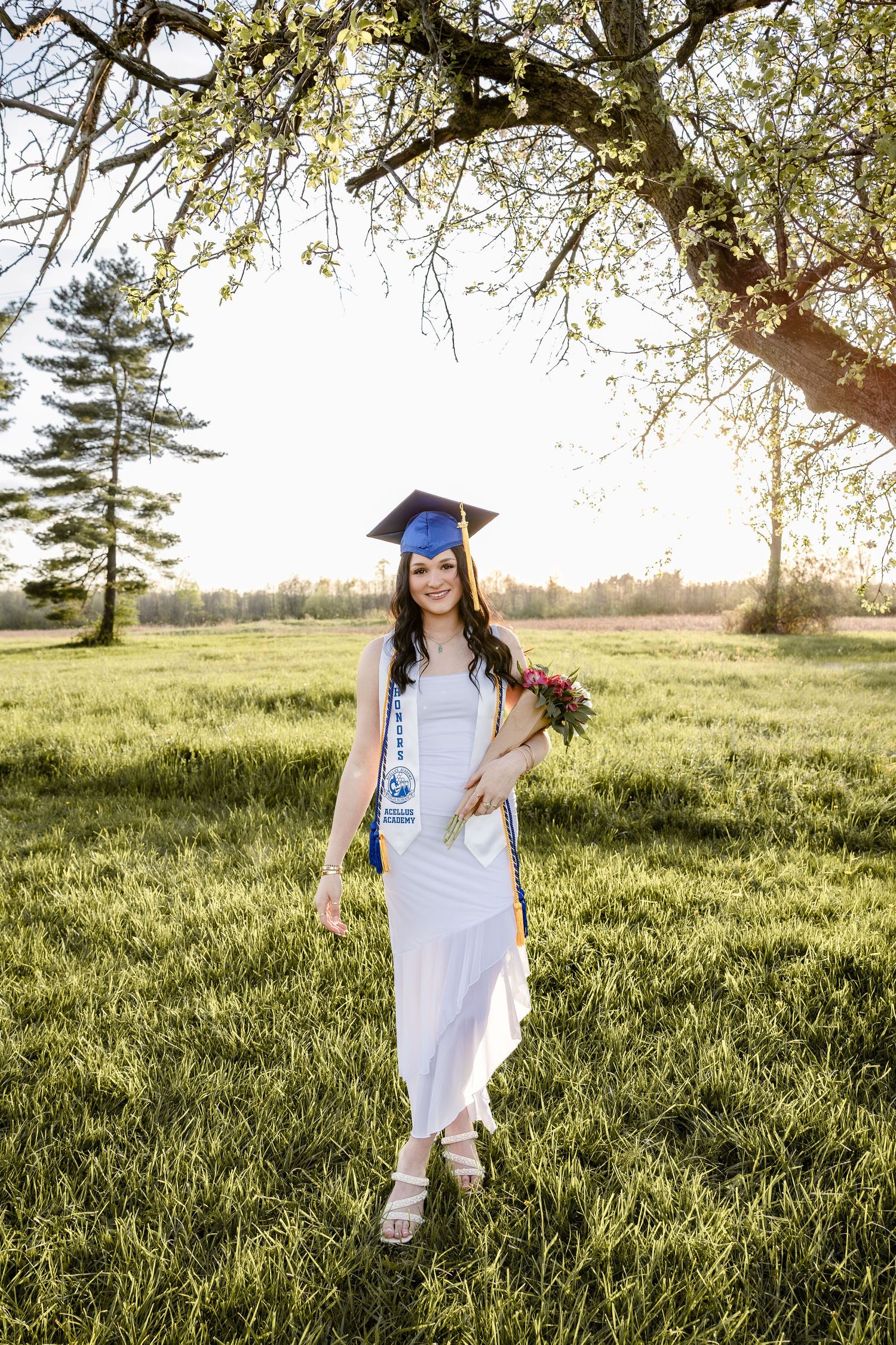 Young woman in blue graduation cap and white gown holding flowers, standing in sunny green field near blooming tree.