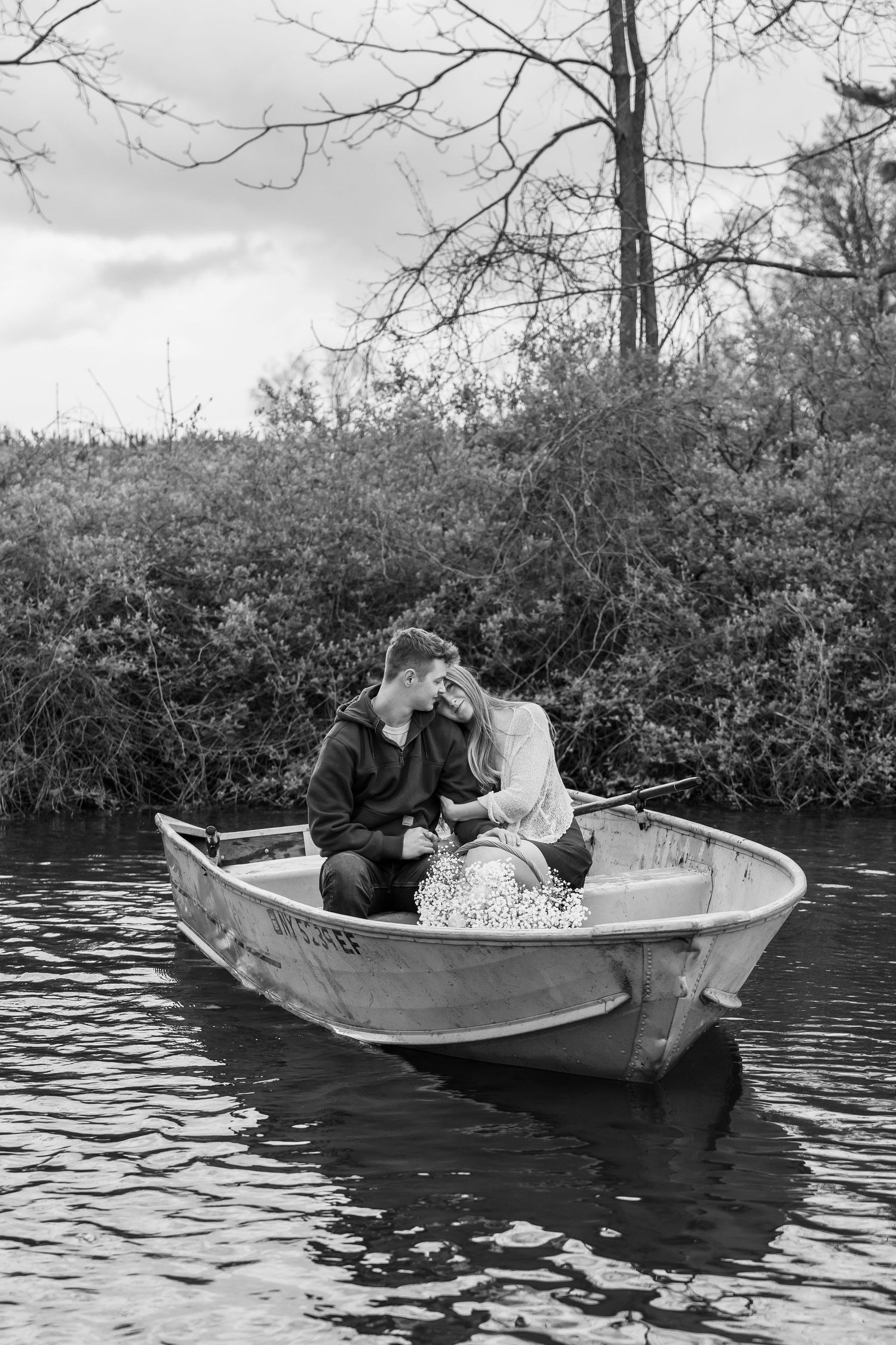 Black and white photo of a romantic couple sitting close together in a small rowboat on a calm river surrounded by trees.