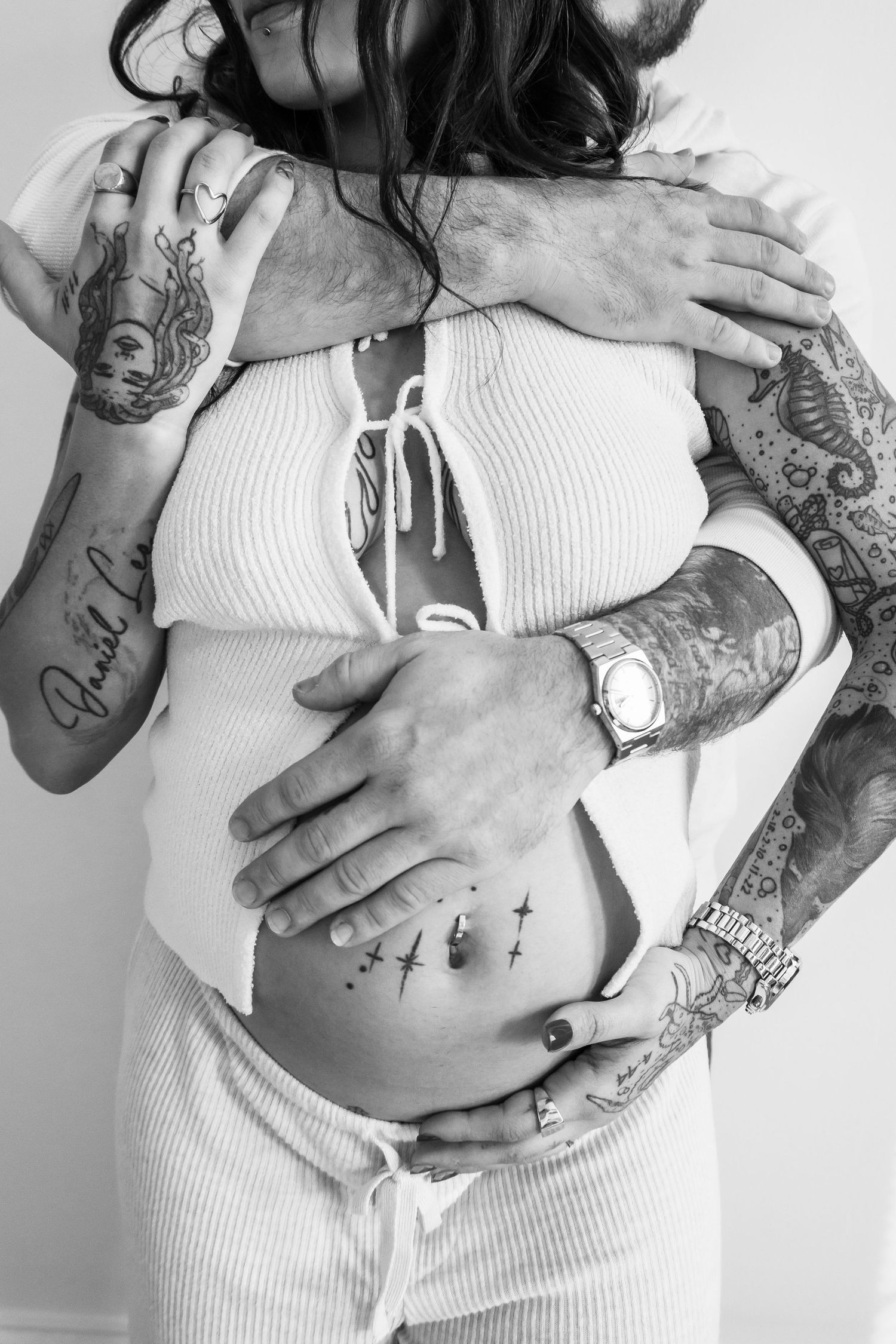 Black and white maternity photo of tattooed couple embracing, man's hands cradling pregnant woman's belly.