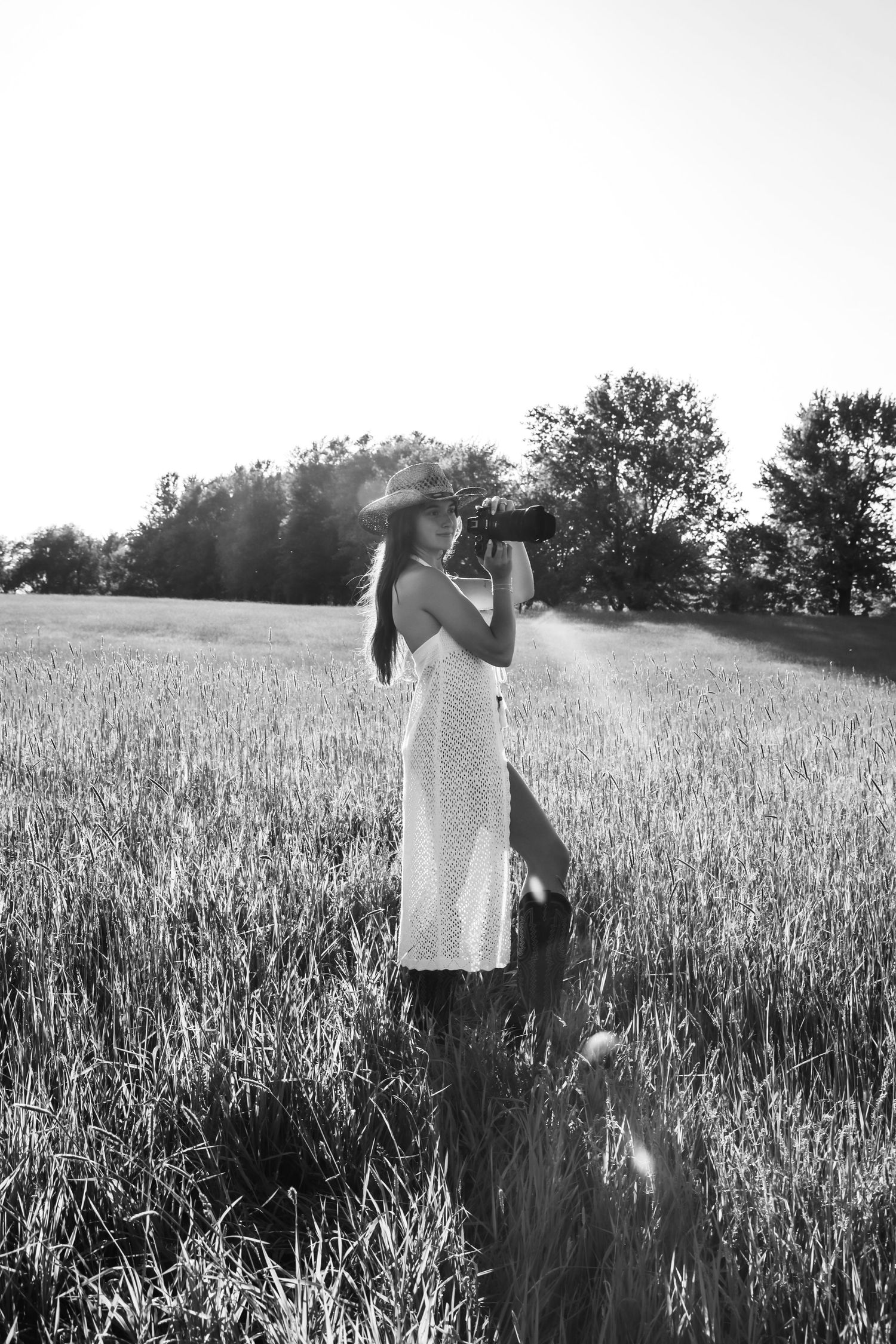 Black and white photo of a woman in a white dress and hat photographing in a sunlit wheat field.