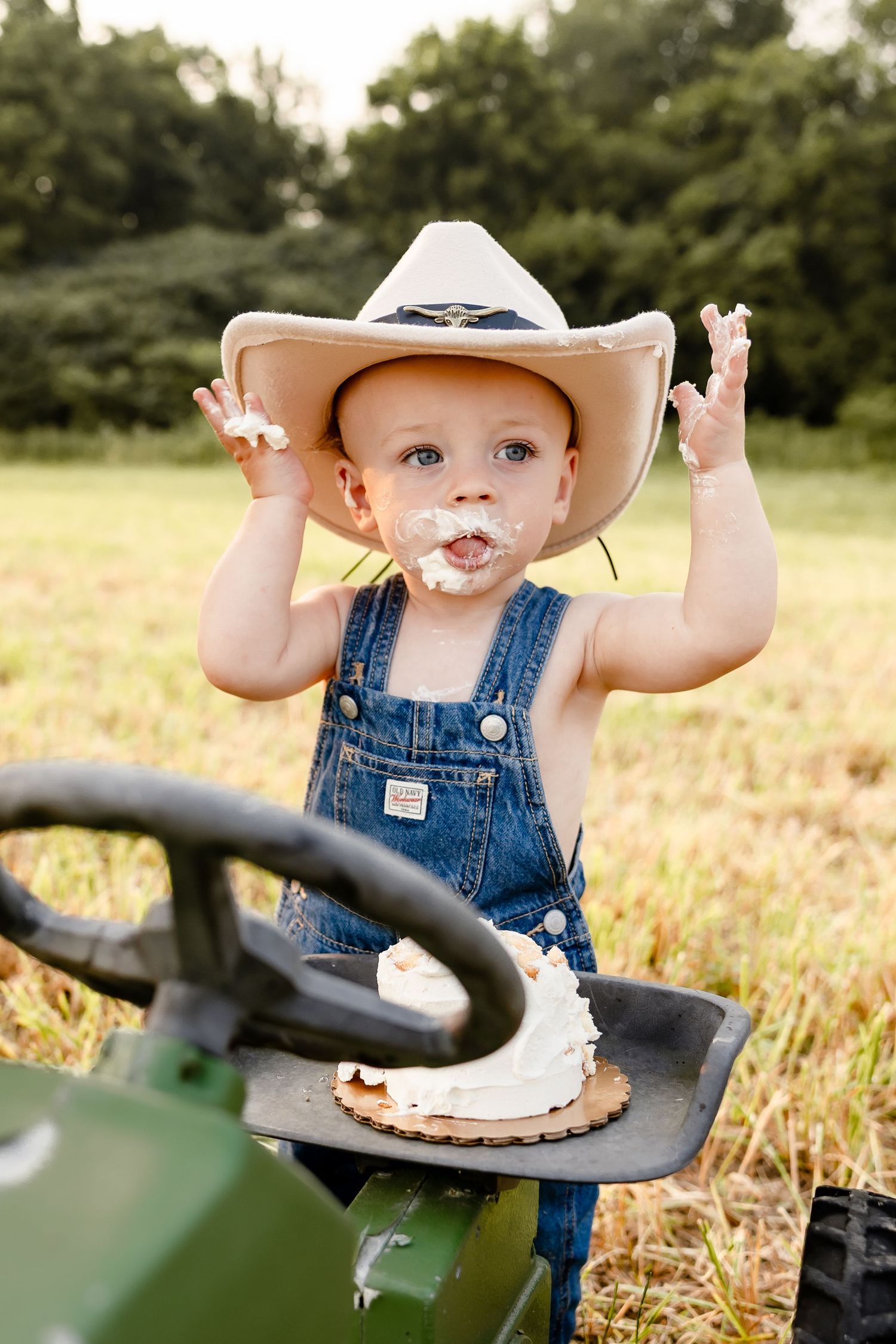 Toddler in denim overalls and cowboy hat smashing birthday cake on green tractor in sunny field.