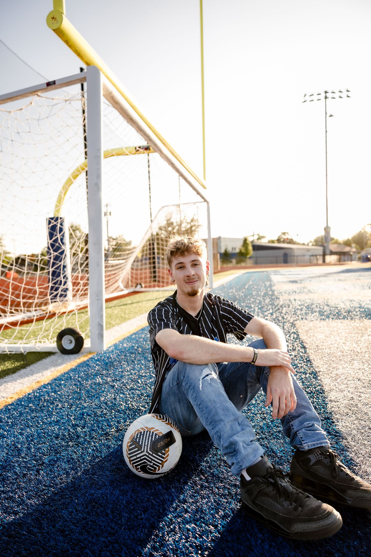 Teen boy sits on blue turf beside a soccer ball and goal post on a sunny day, wearing jeans and a striped shirt.