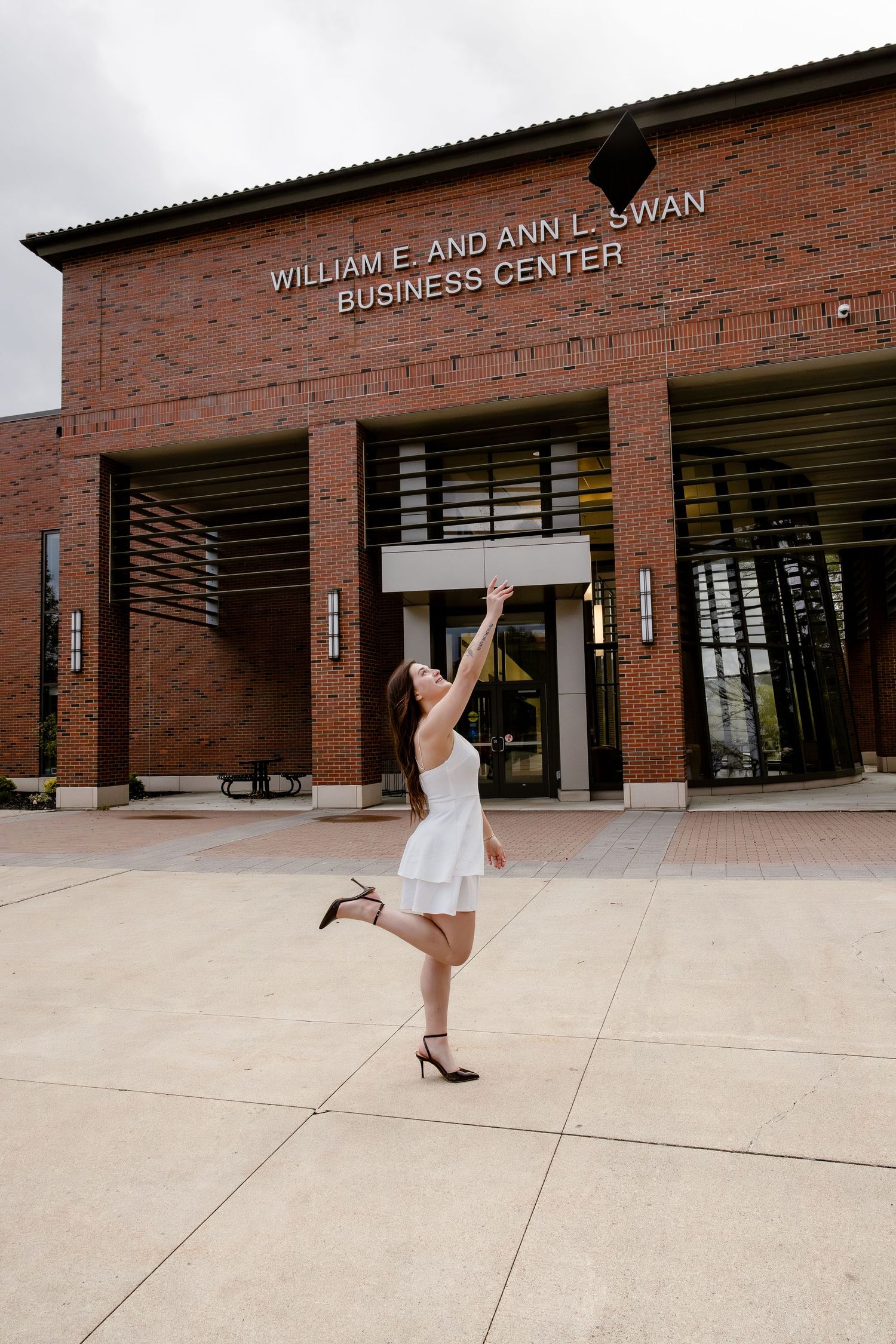 Graduate in white dress joyfully tosses cap outside William E. and Ann L. Swan Business Center.