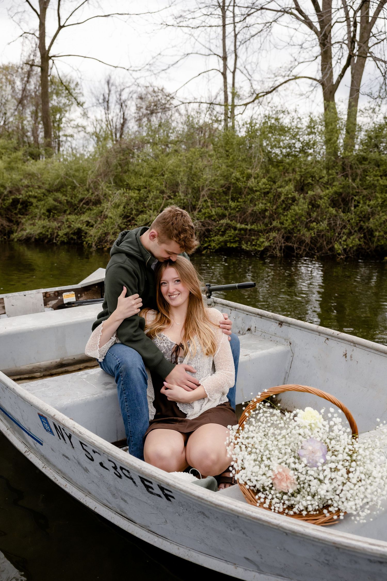 Romantic couple embracing in a white rowboat on a calm river, surrounded by trees, with a basket of white flowers.