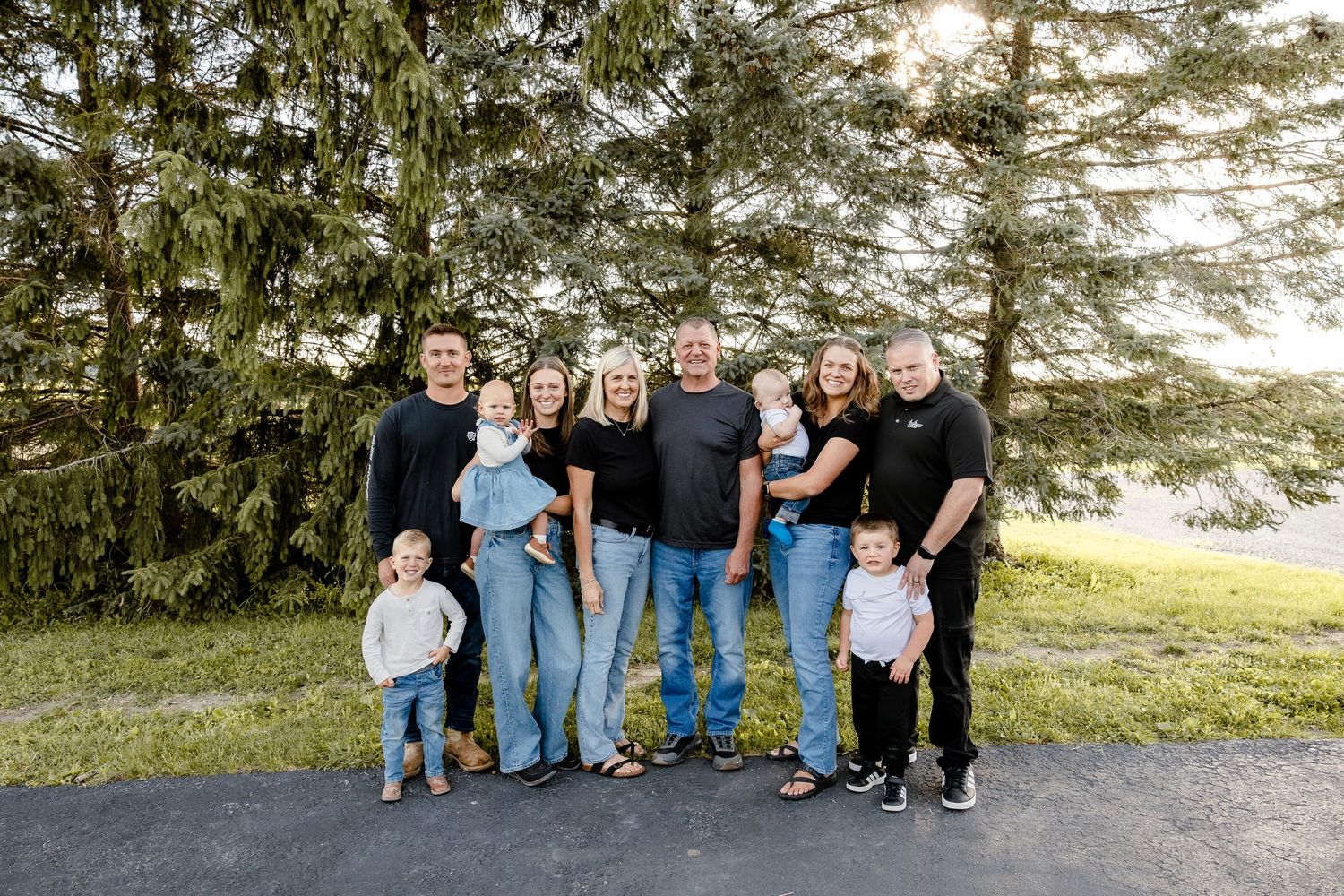 Large family group portrait outdoors near tall pine trees on a sunny day, smiling adults and young children on a driveway.