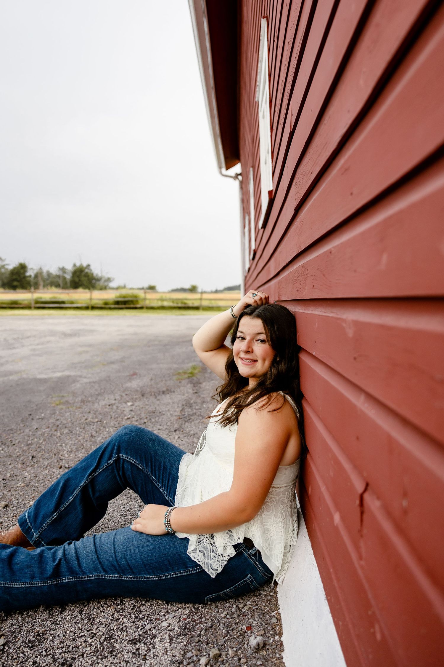 Young woman in jeans and white top sitting against a red barn wall, smiling outdoors on a gravel path.