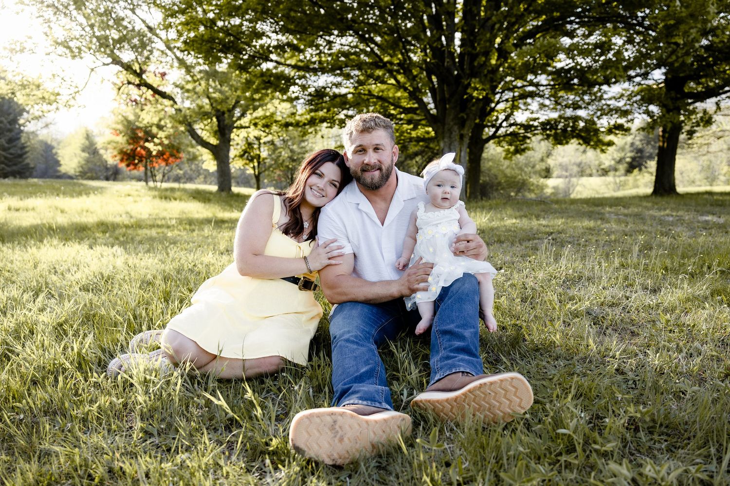 Family portrait of couple with baby girl sitting in green meadow under trees in warm golden sunlight.
