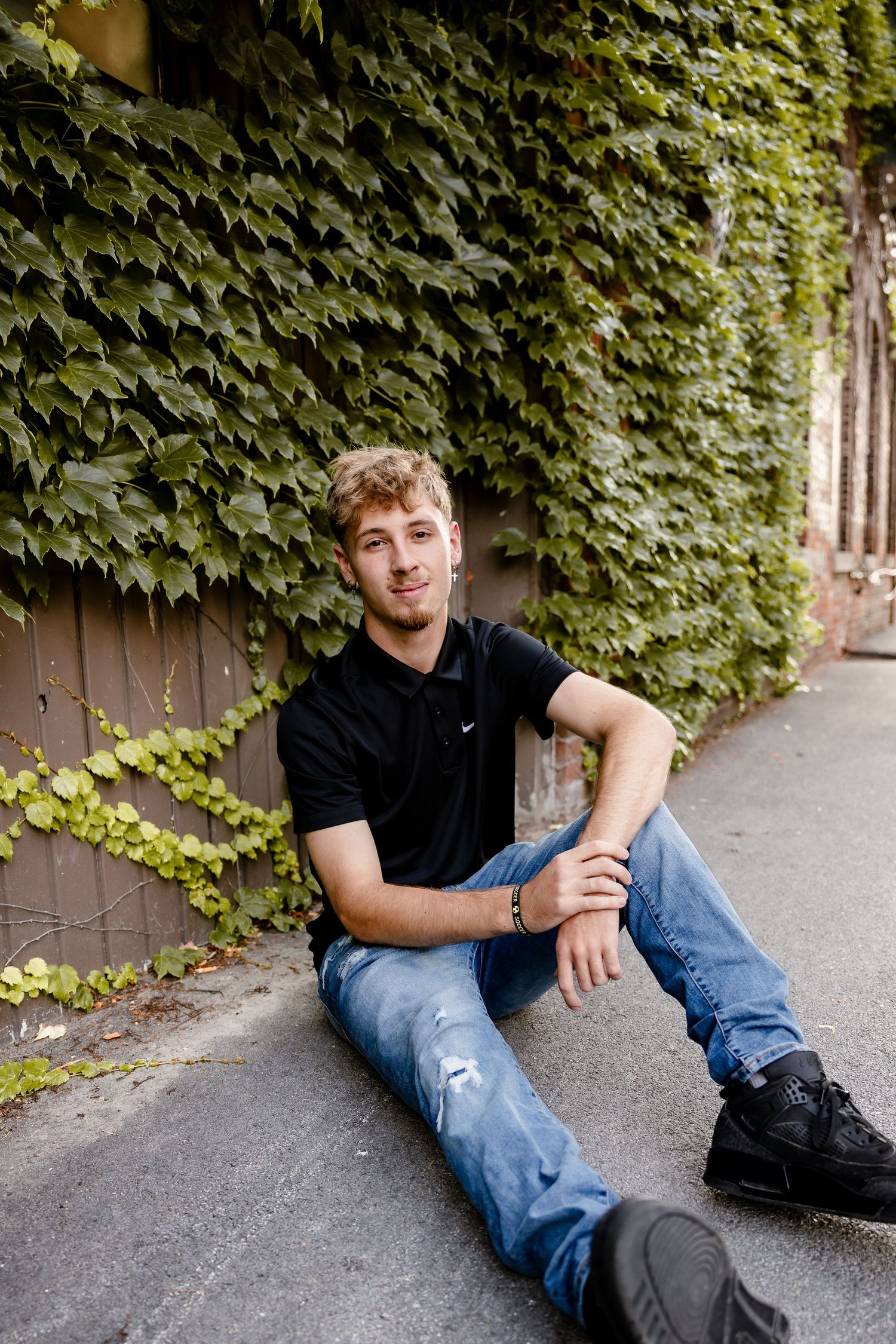 Young man in black Nike polo and ripped jeans sitting against ivy-covered wall on urban sidewalk.