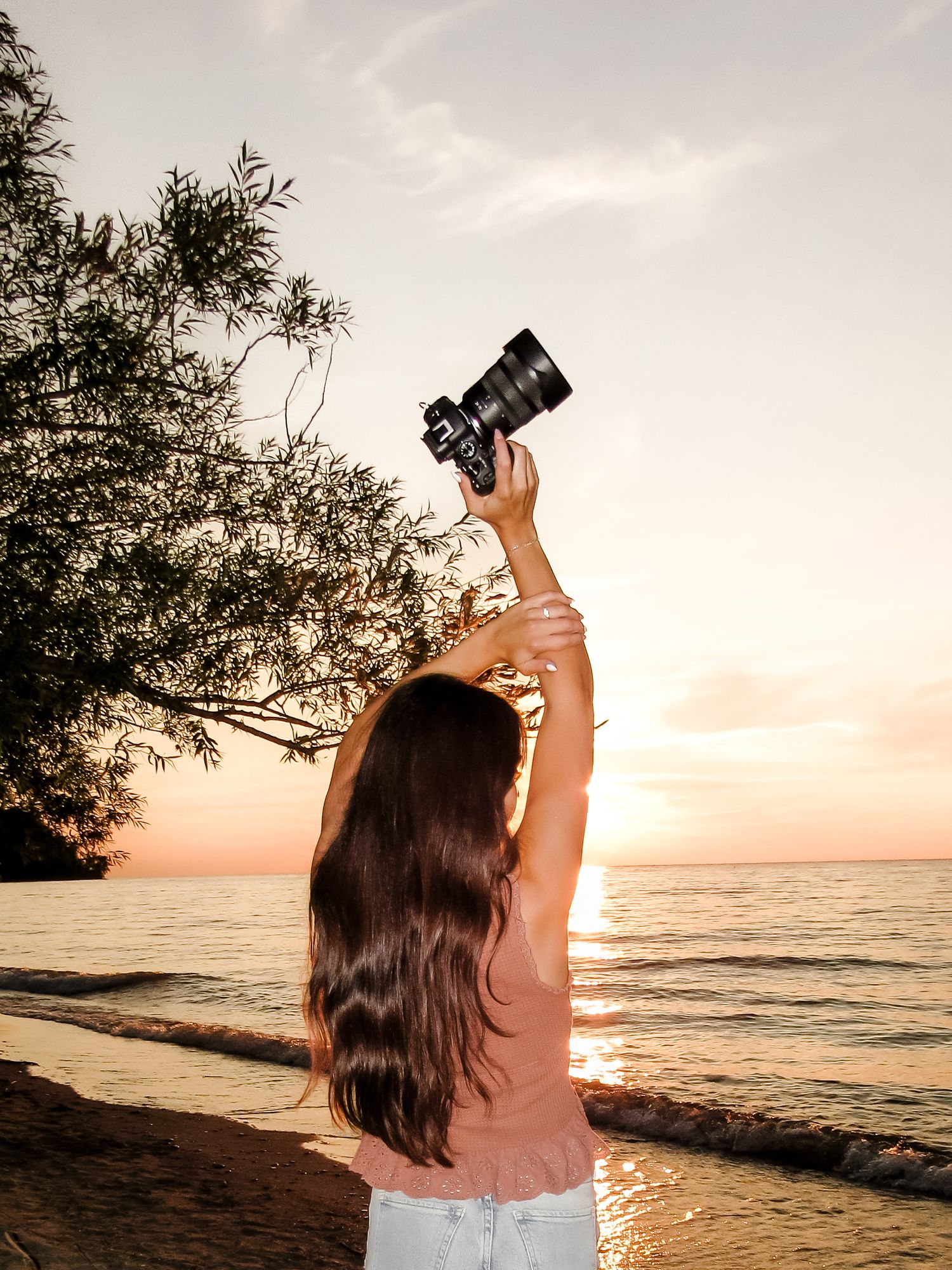 Woman holding camera overhead at golden hour beach sunset with warm orange sky and calm ocean waters.