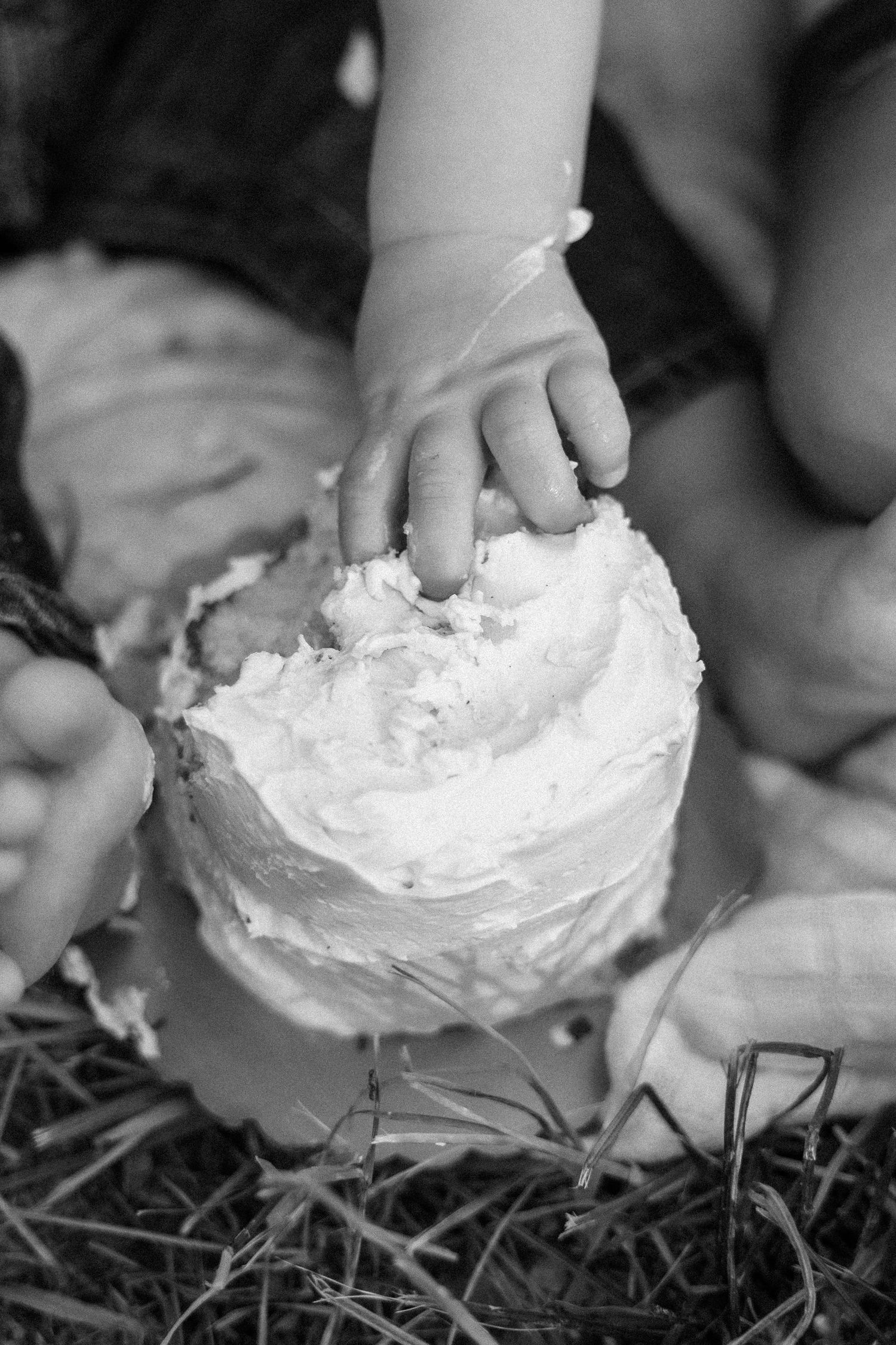 Black and white photo of a baby's hand touching a frosted smash cake outdoors on grass.