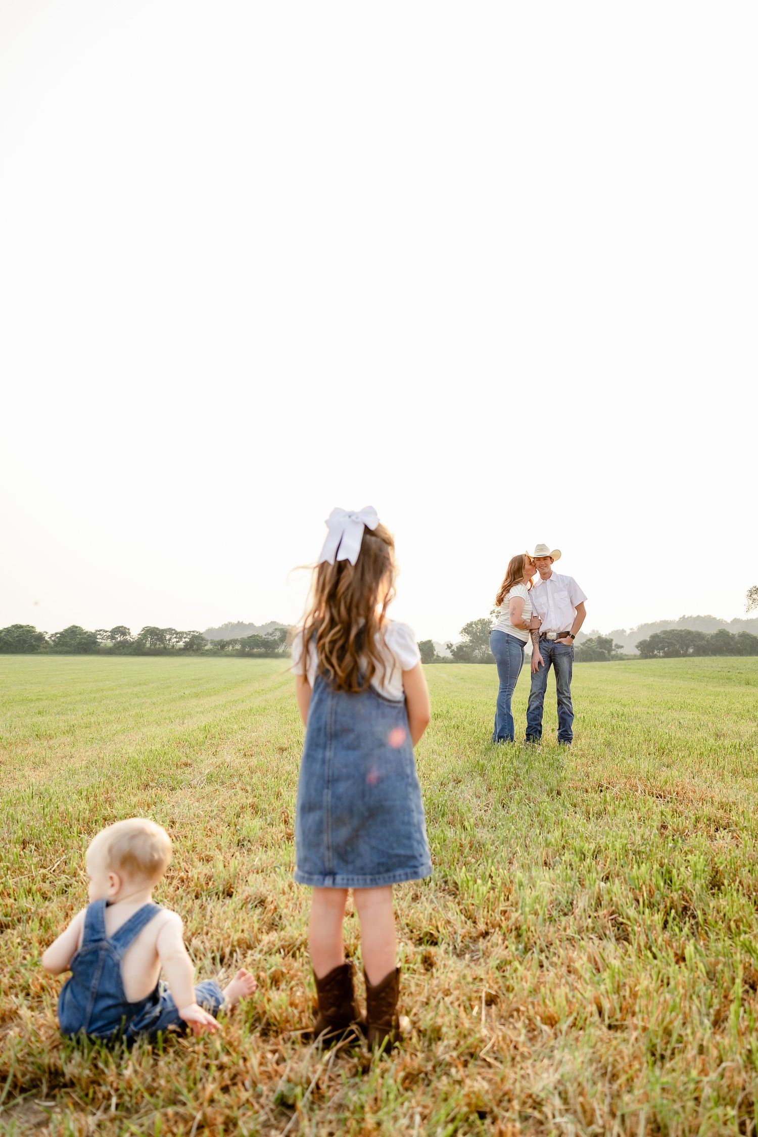 Family in a green field at golden hour, two children in denim and a couple embracing in the background.