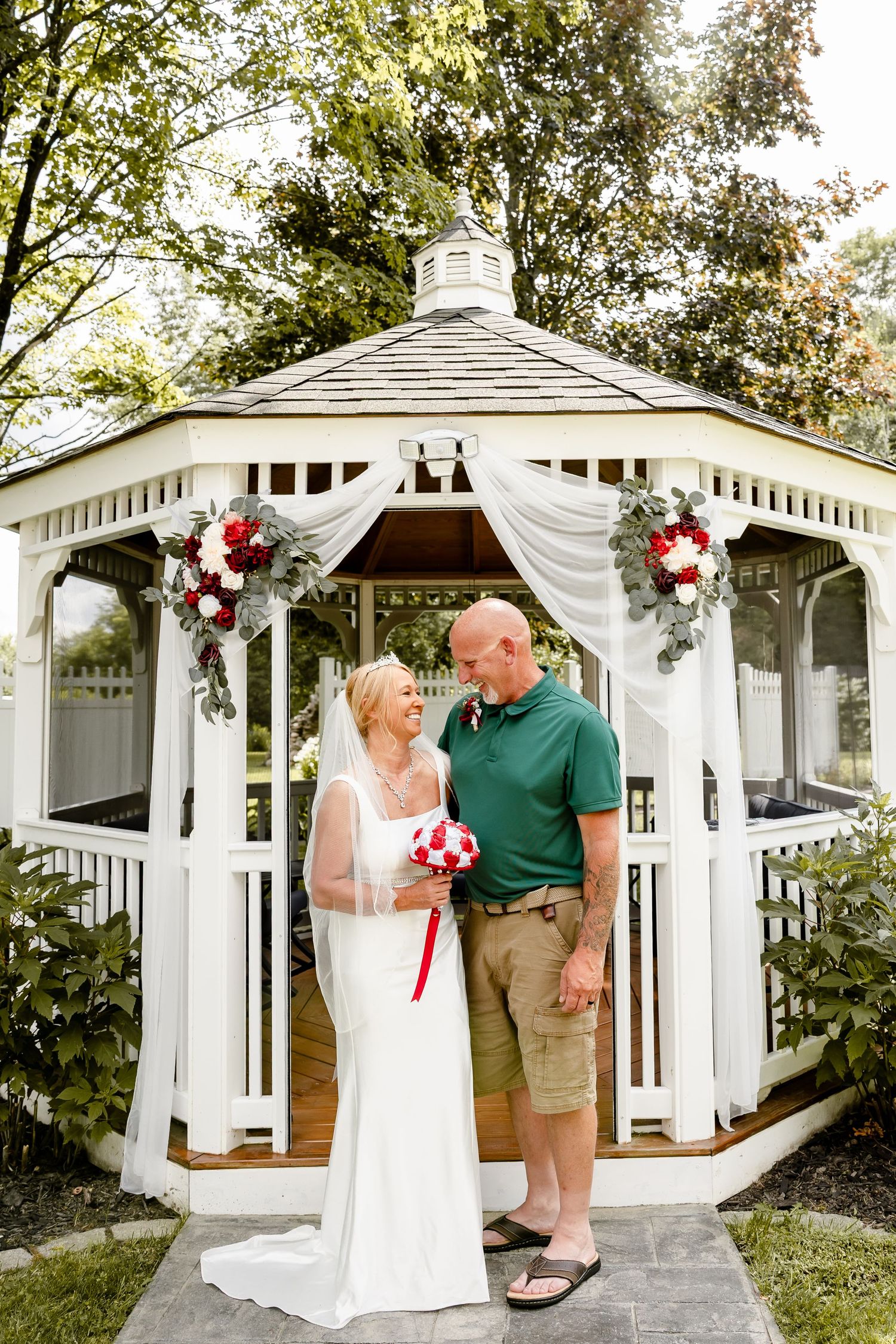 Bride and groom smiling together in front of a white gazebo decorated with red and white floral arrangements.