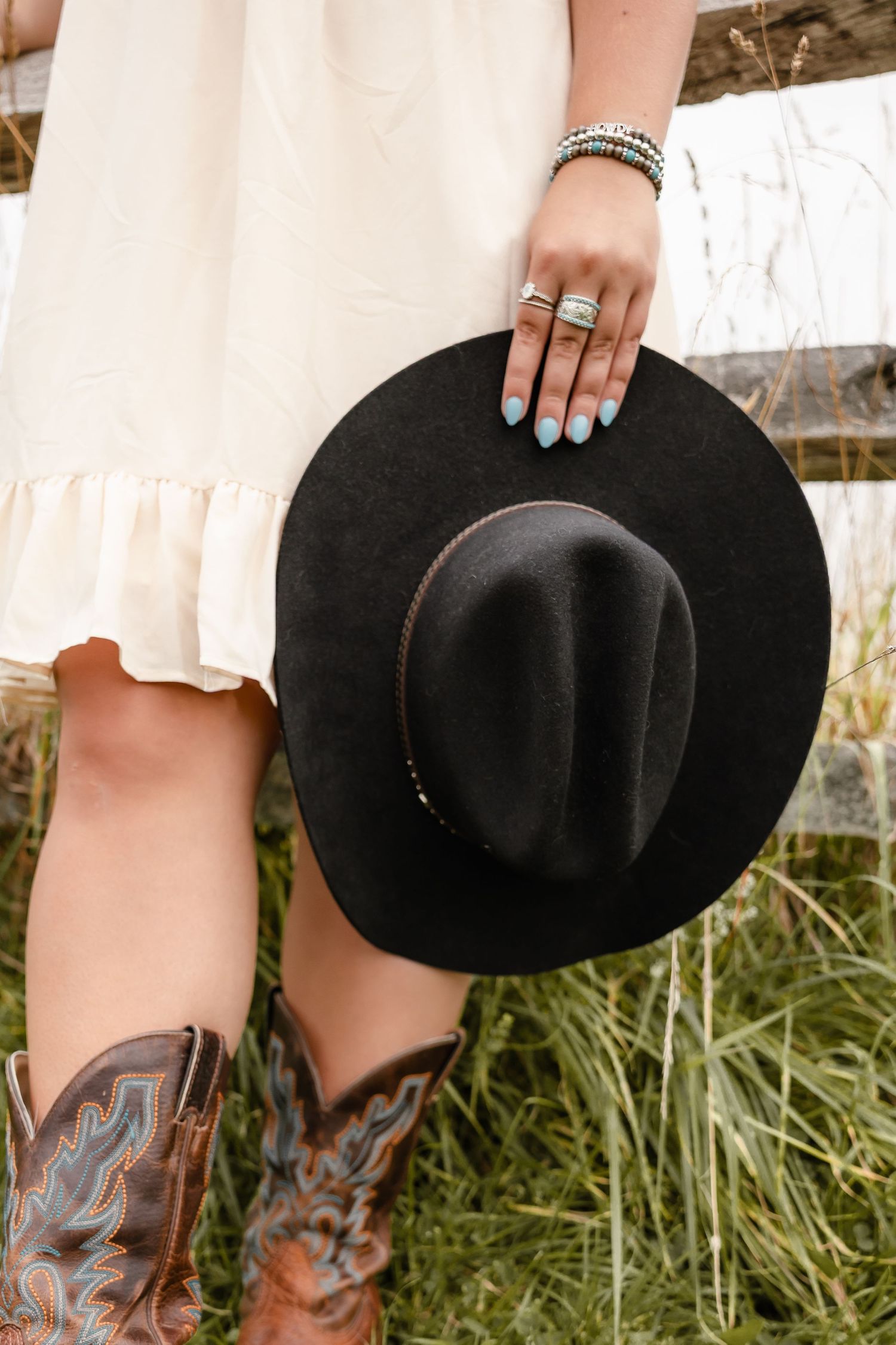 Woman in white dress and cowboy boots holding a black felt wide-brim hat with silver rings and turquoise nails.