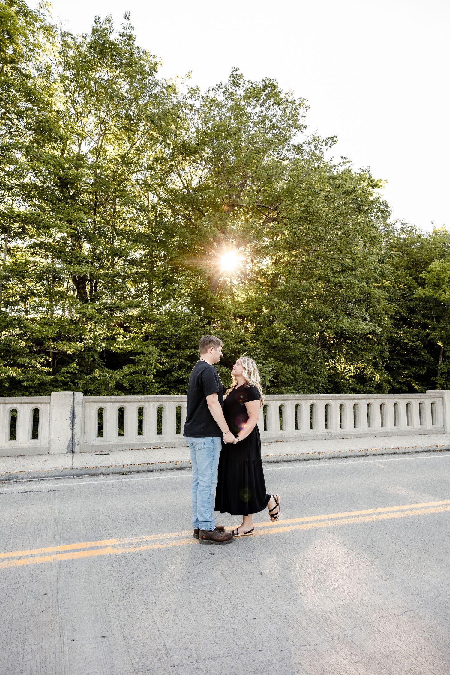 Couple holding hands on a bridge at golden hour, surrounded by lush green trees with sunlight streaming through.