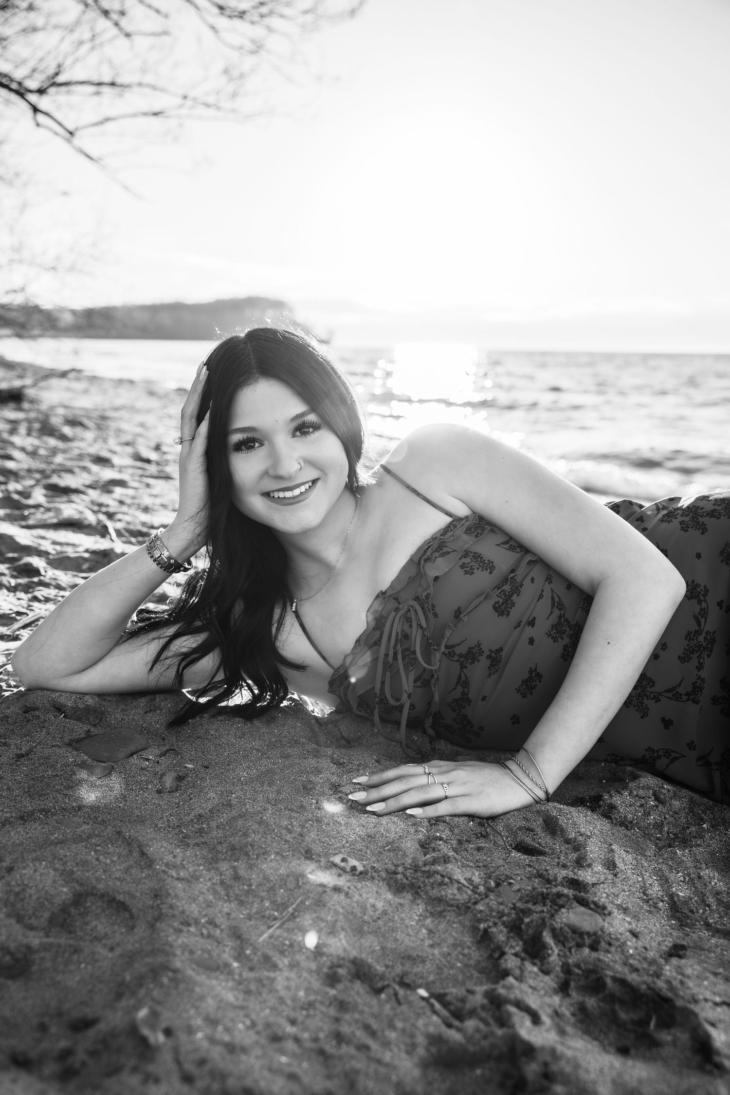 Black and white photo of a smiling woman lying on a rocky beach with ocean and sunset in the background.