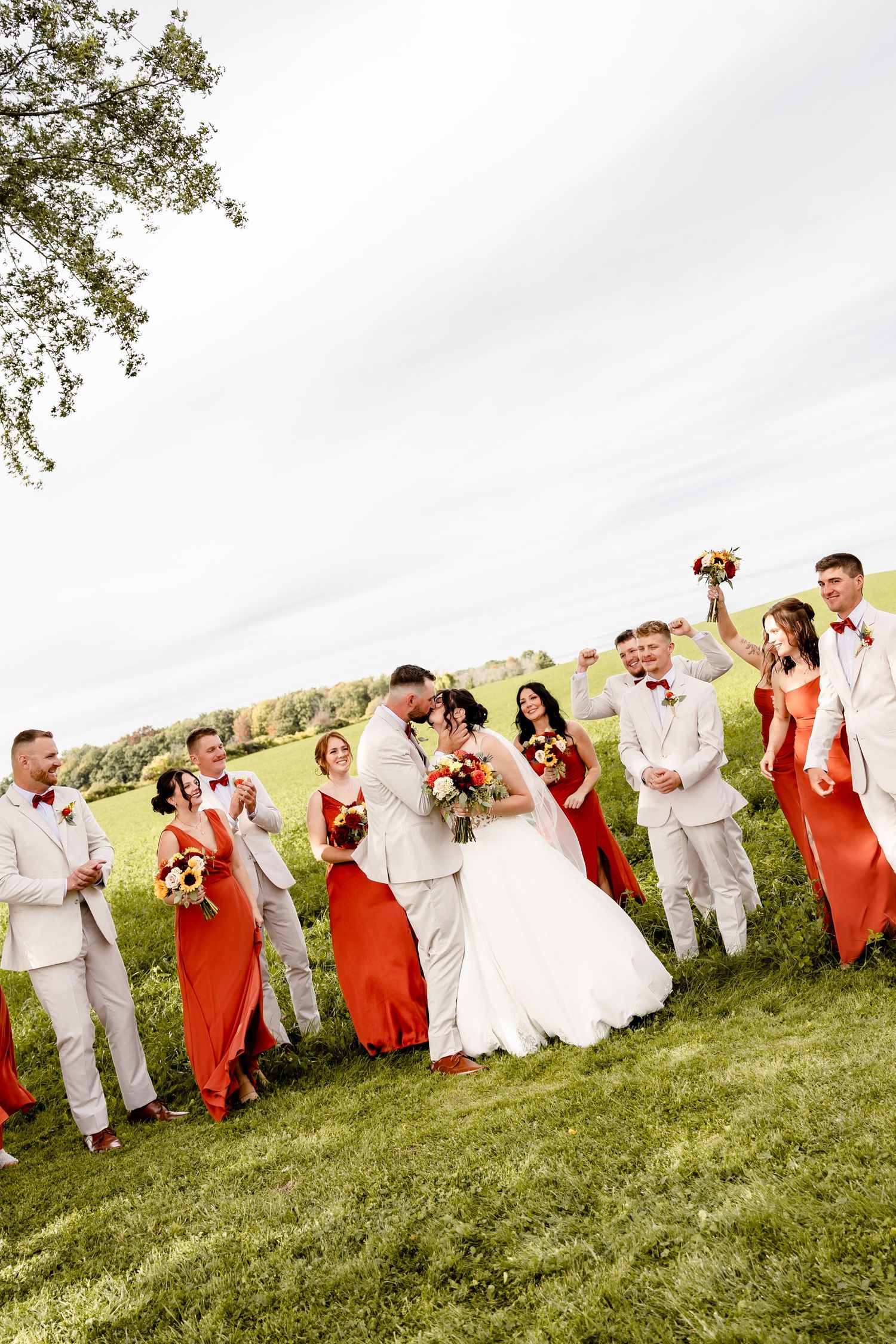 Wedding party in red dresses and tan suits celebrate with bride and groom kissing outdoors on a green lawn.