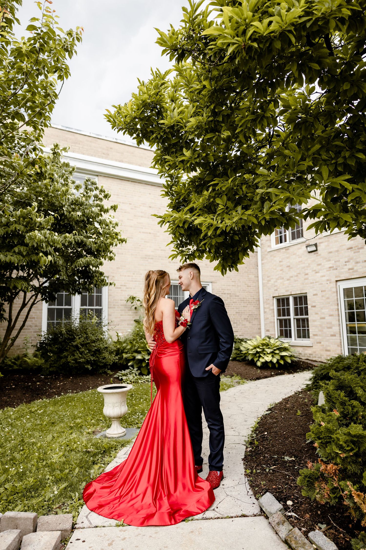 Couple in formal attire, woman in red gown and man in navy suit, posing in a garden courtyard.