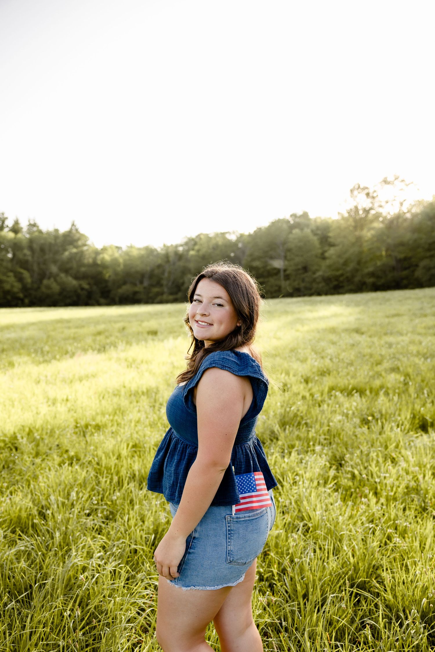 Young woman in denim outfit with American flag pocket stands smiling in a sunlit green field.
