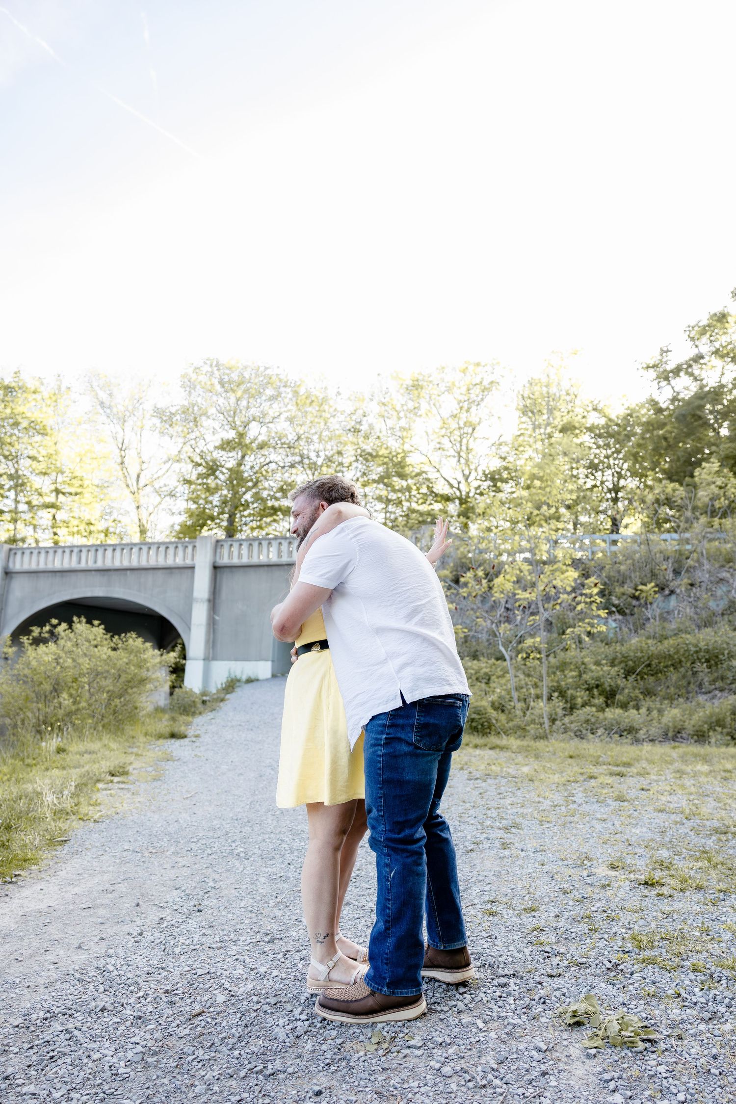 Couple embracing on gravel path near stone bridge, woman in yellow dress and man in white shirt and jeans sharing a hug.