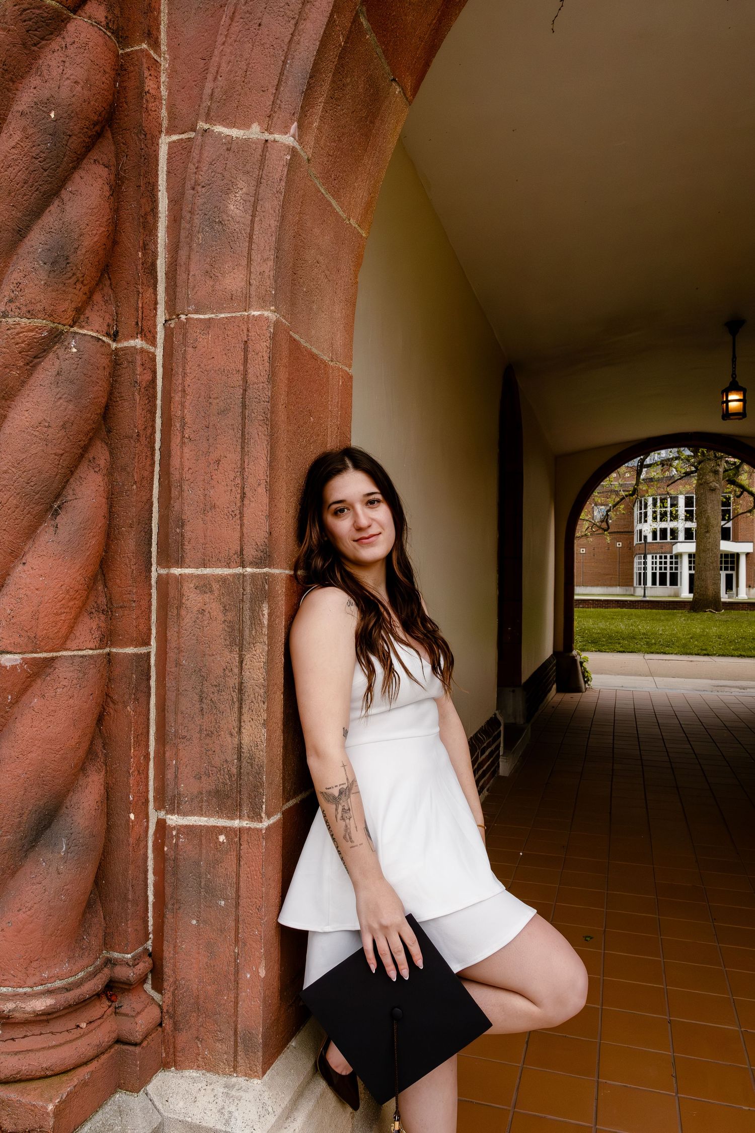 Young woman in white dress holding graduation cap, posing by ornate brick archway on college campus.