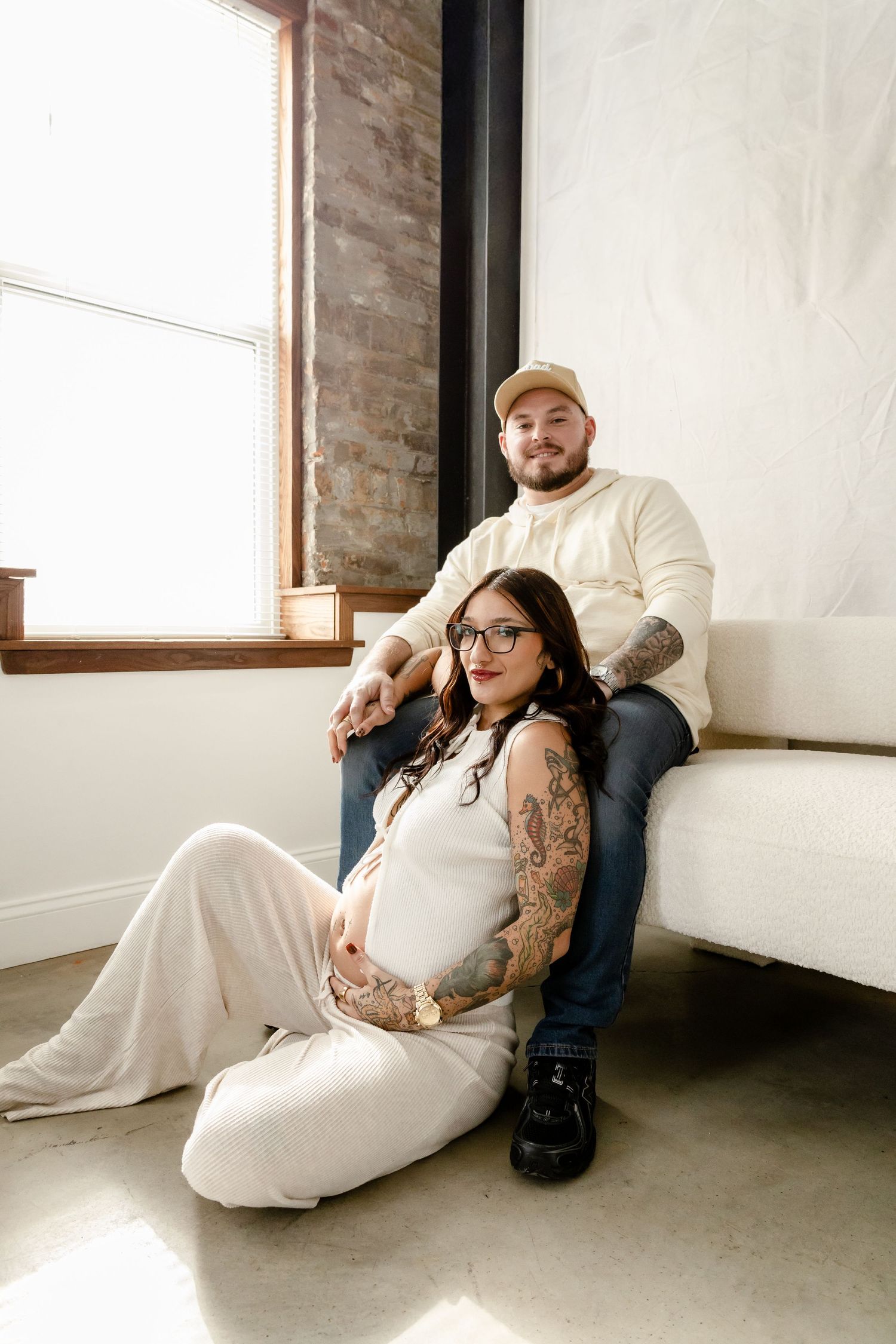 Tattooed pregnant couple posing together, woman in white jumpsuit sitting on floor, man in cream sweater on couch.