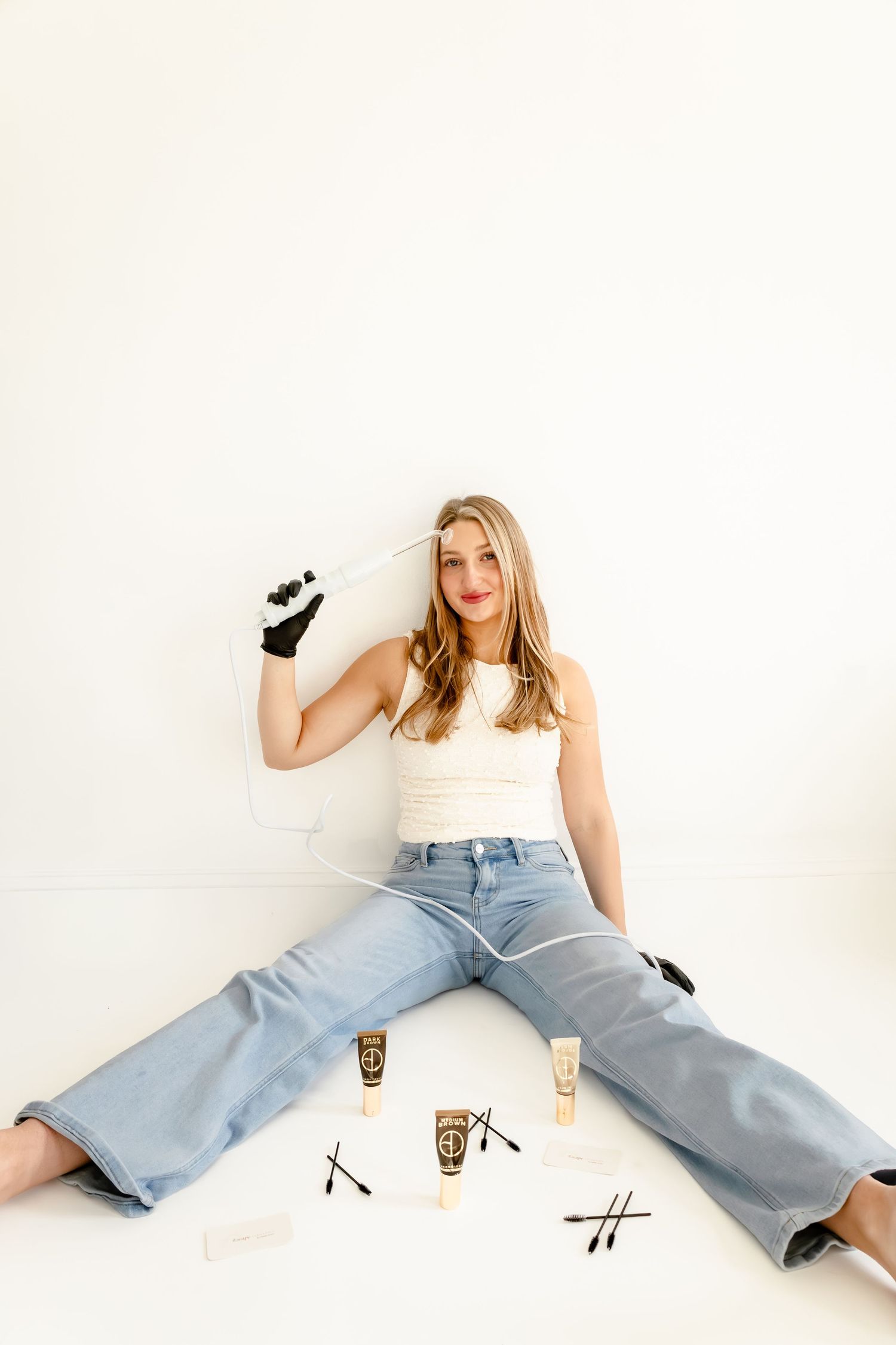 Woman in white tank and jeans sits on floor with makeup tubes and brushes, holding applicator tool against white wall.