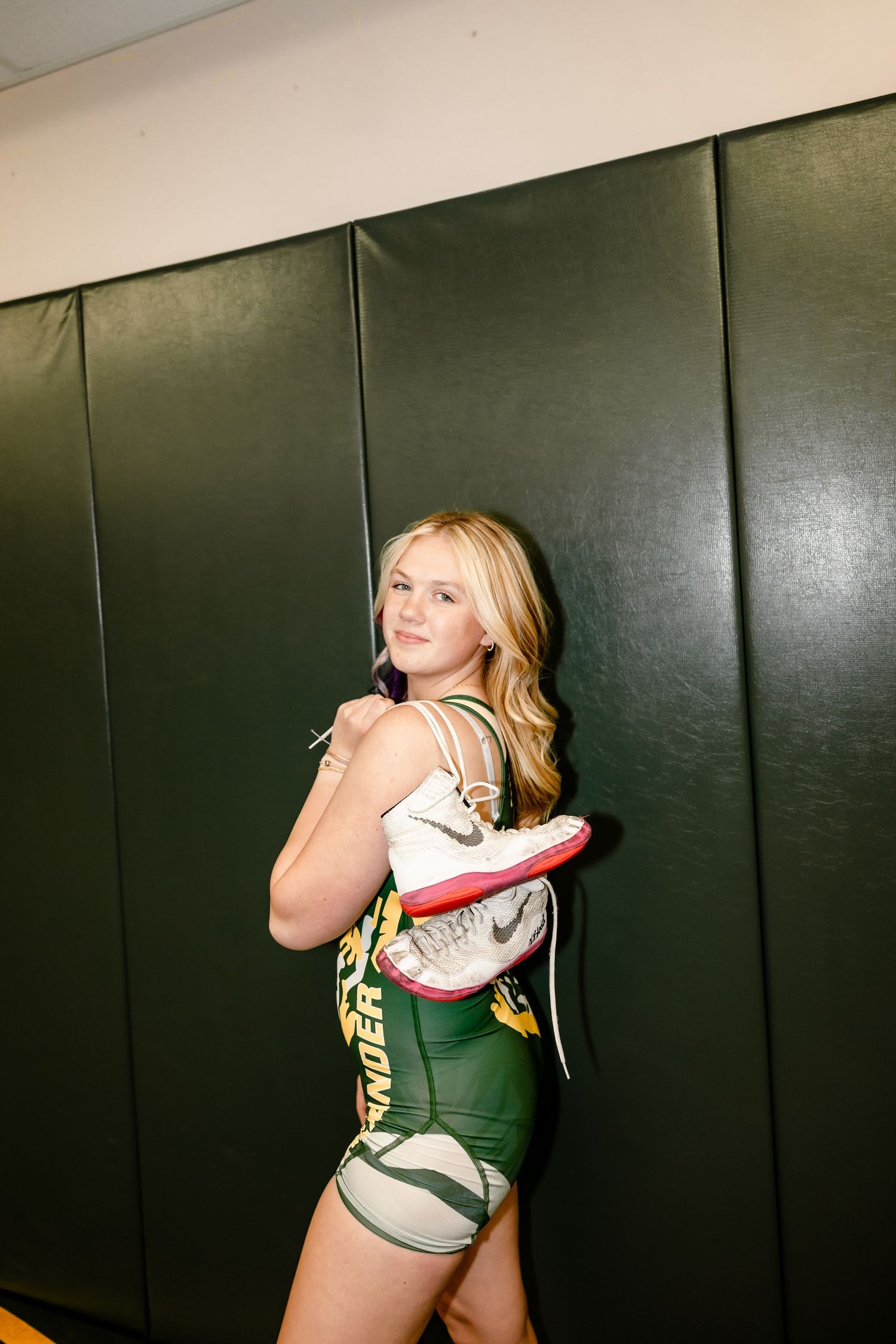 Female wrestler in green singlet holds athletic shoes over shoulder, posing confidently against green gym wall padding.