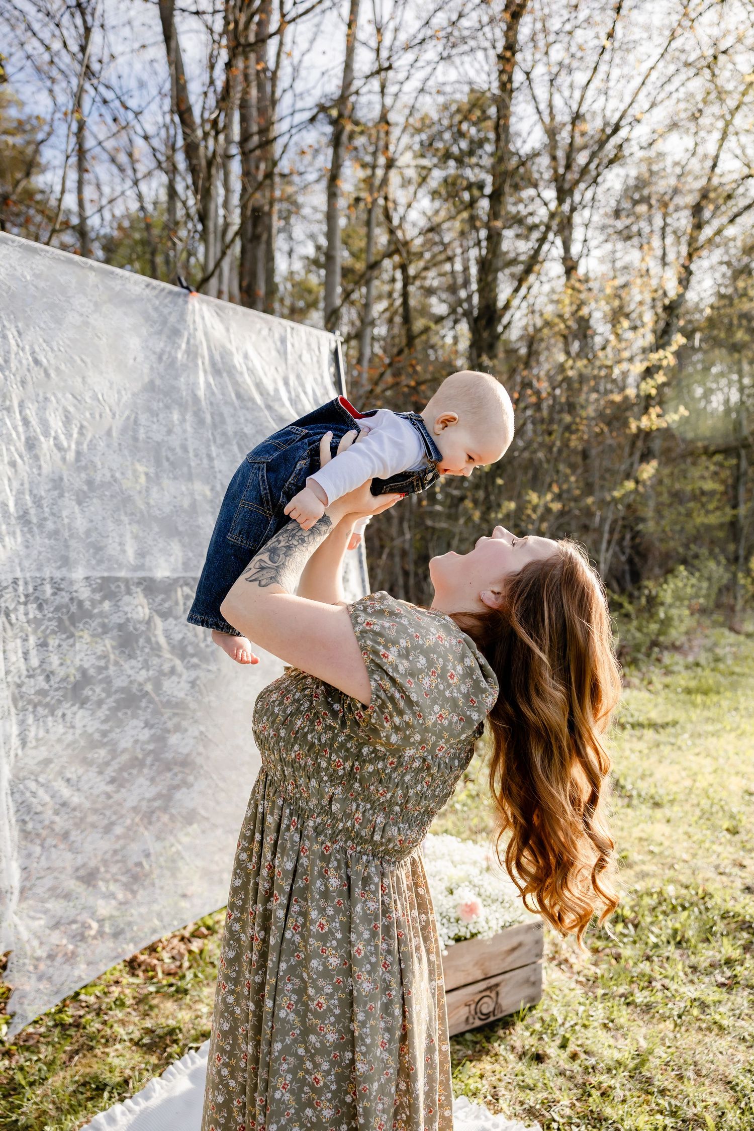A mother in a floral dress lifts her baby joyfully outdoors near trees and a white backdrop.