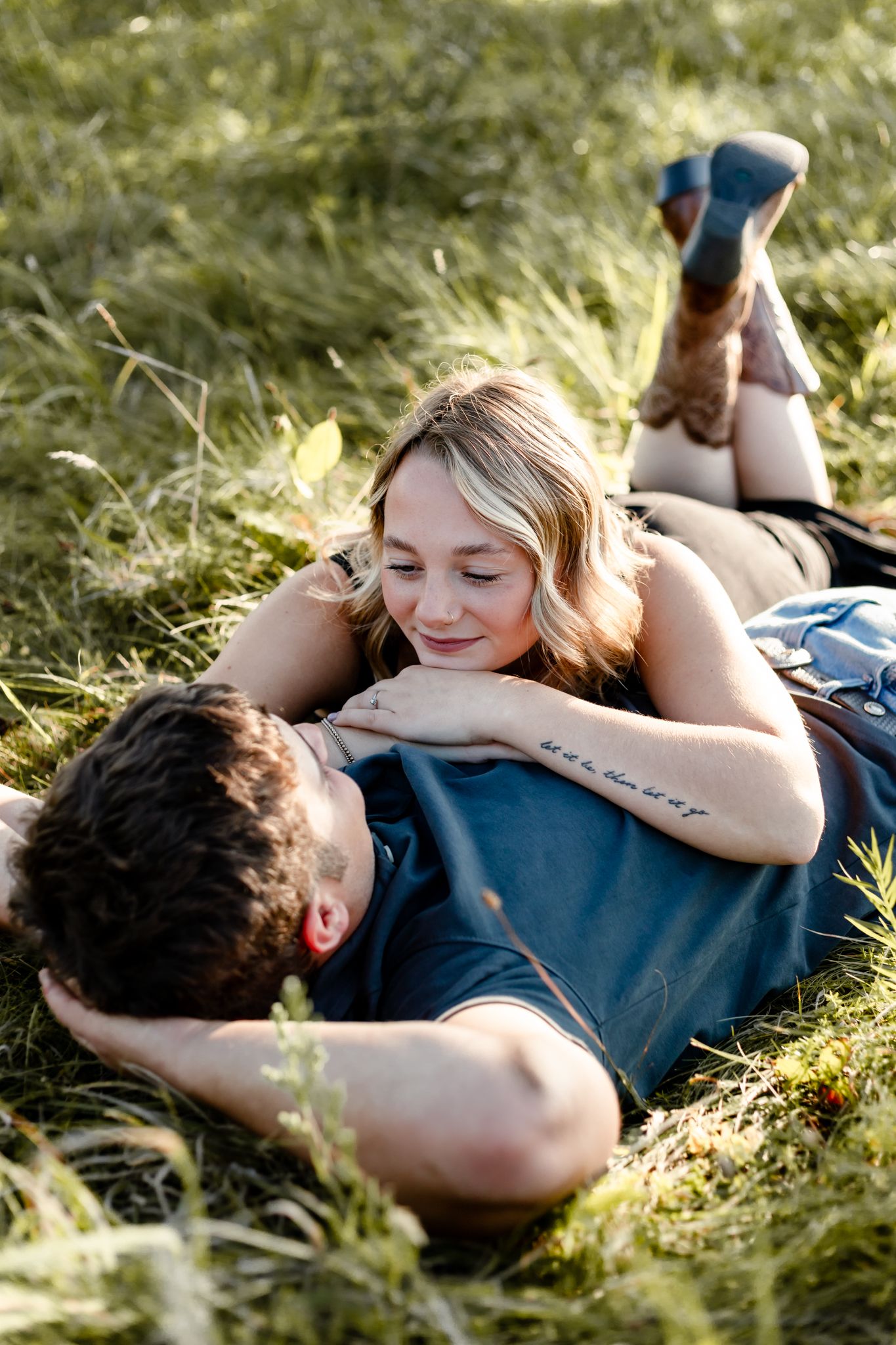 A couple relaxes in green grass, woman with tattoo resting on man's chest, smiling softly in warm sunlight.