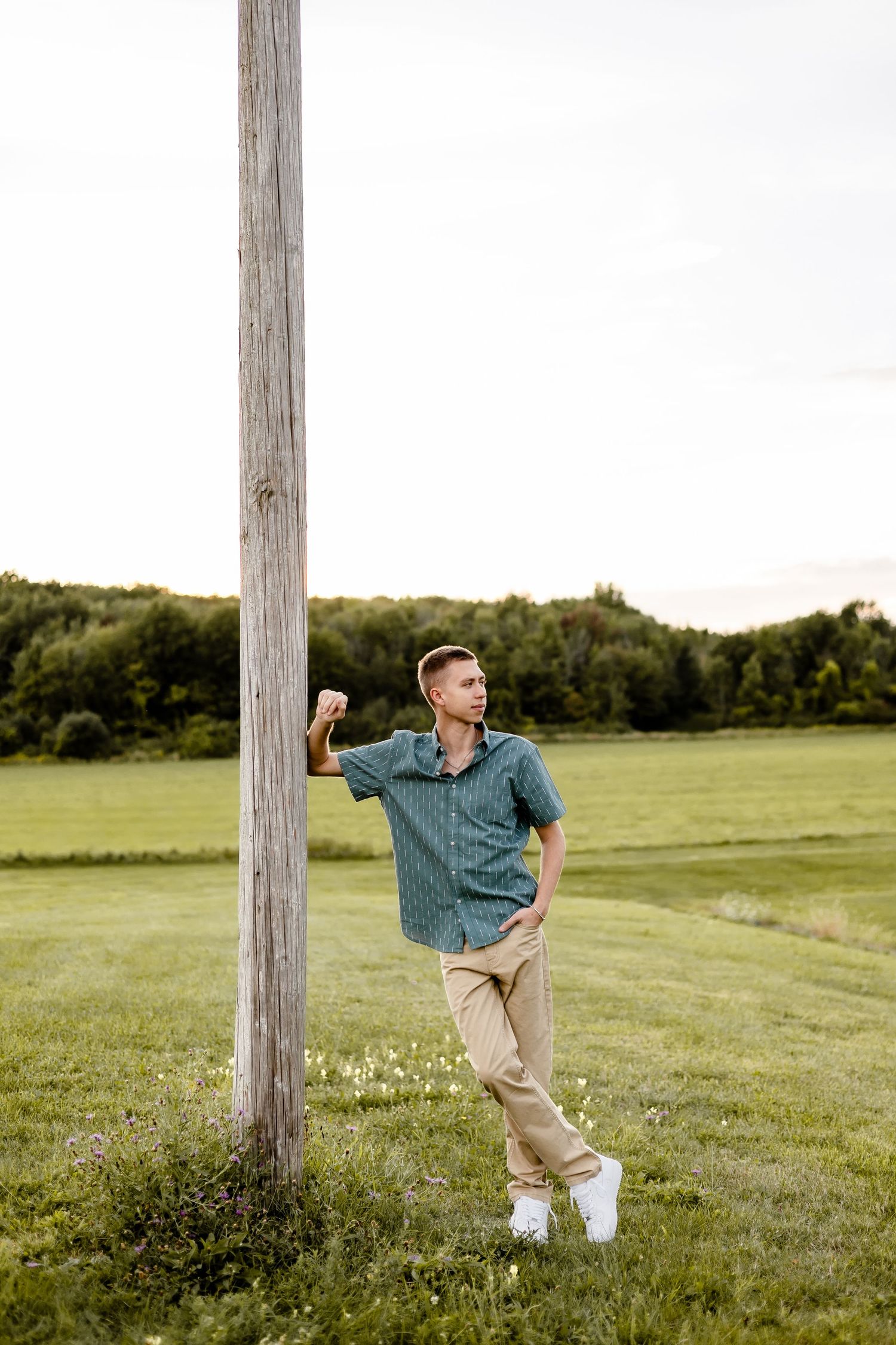 Young man in green shirt and khaki pants leaning against a wooden pole in a lush green field.
